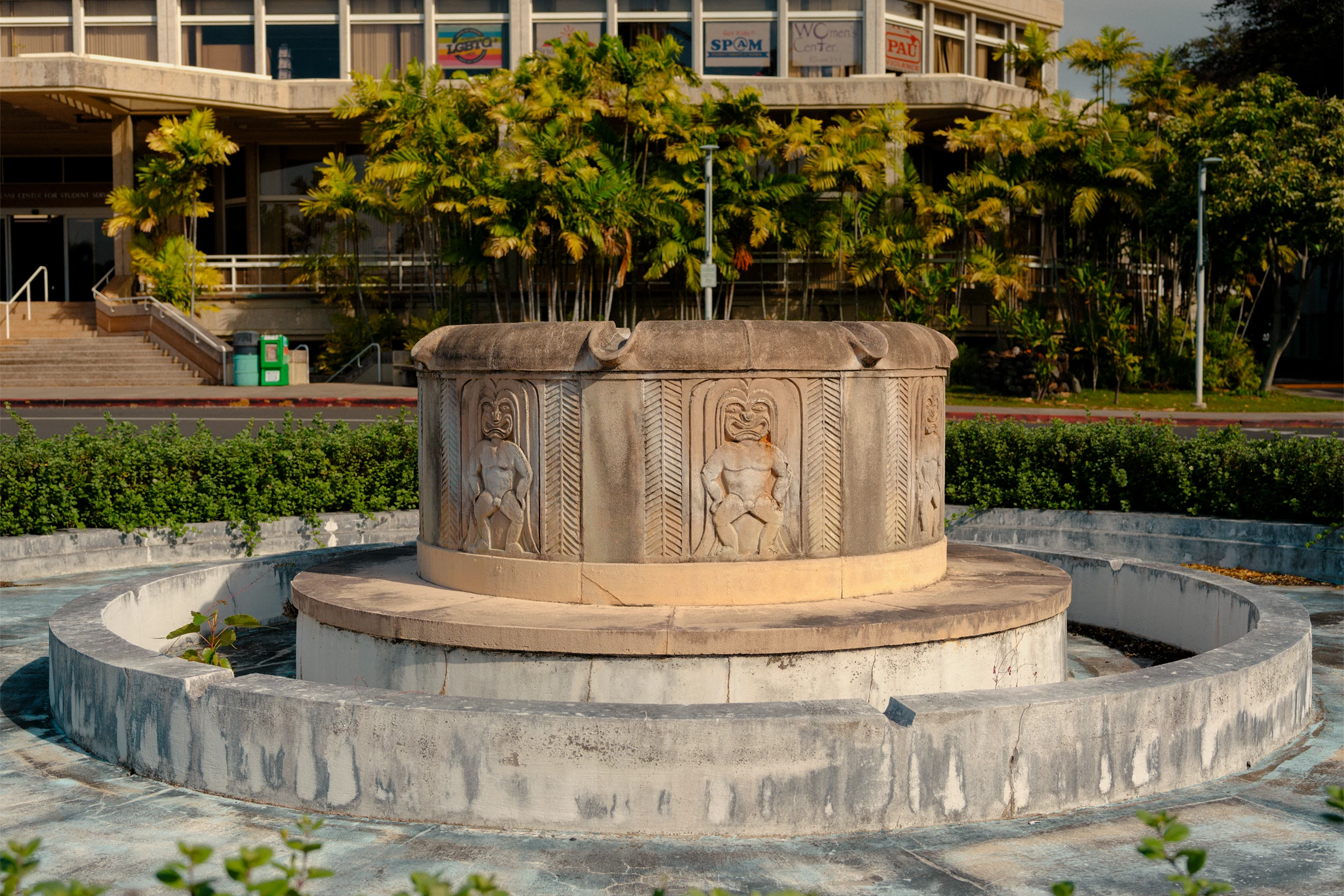 An empty round stone fountain with carved native Hawai'an designs on its sides among greenery at the University of Hawai'i at Mānoa