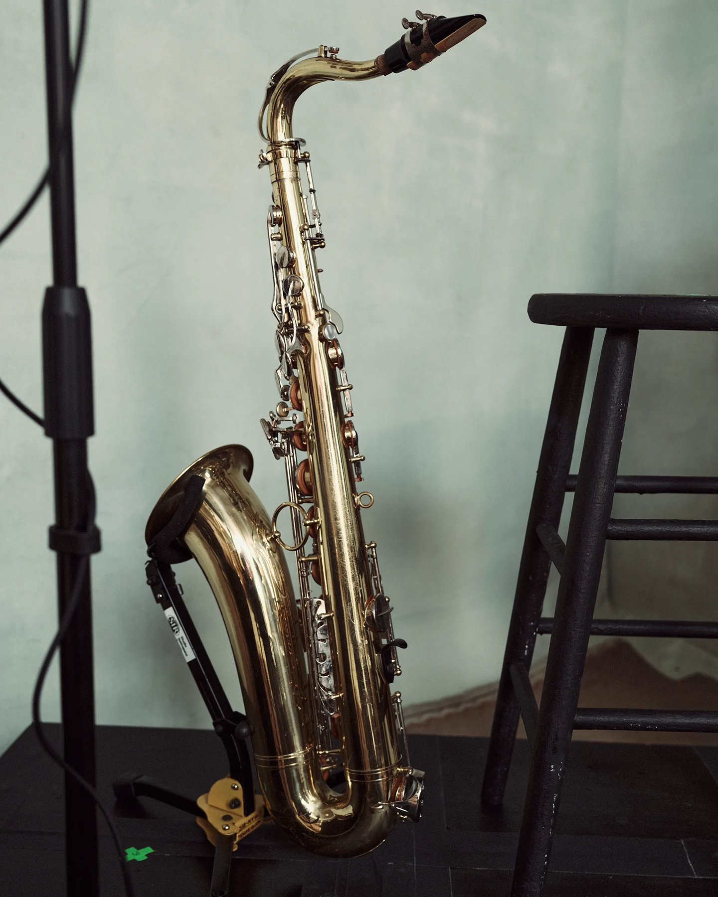 Close-up of a brass tenor saxophone resting on a floor stand beside a black stool and microphone stand. The instrument is set against a soft blue-gray studio backdrop, emphasizing its polished finish and curved shape.