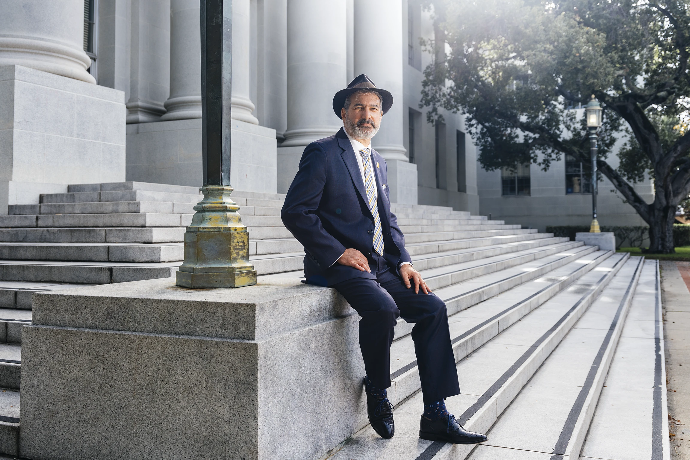A person in a navy suit and gray tie and a fedora hat with a brown and gray beard and black shoes sitting beside a lampost on the steps of a columned building