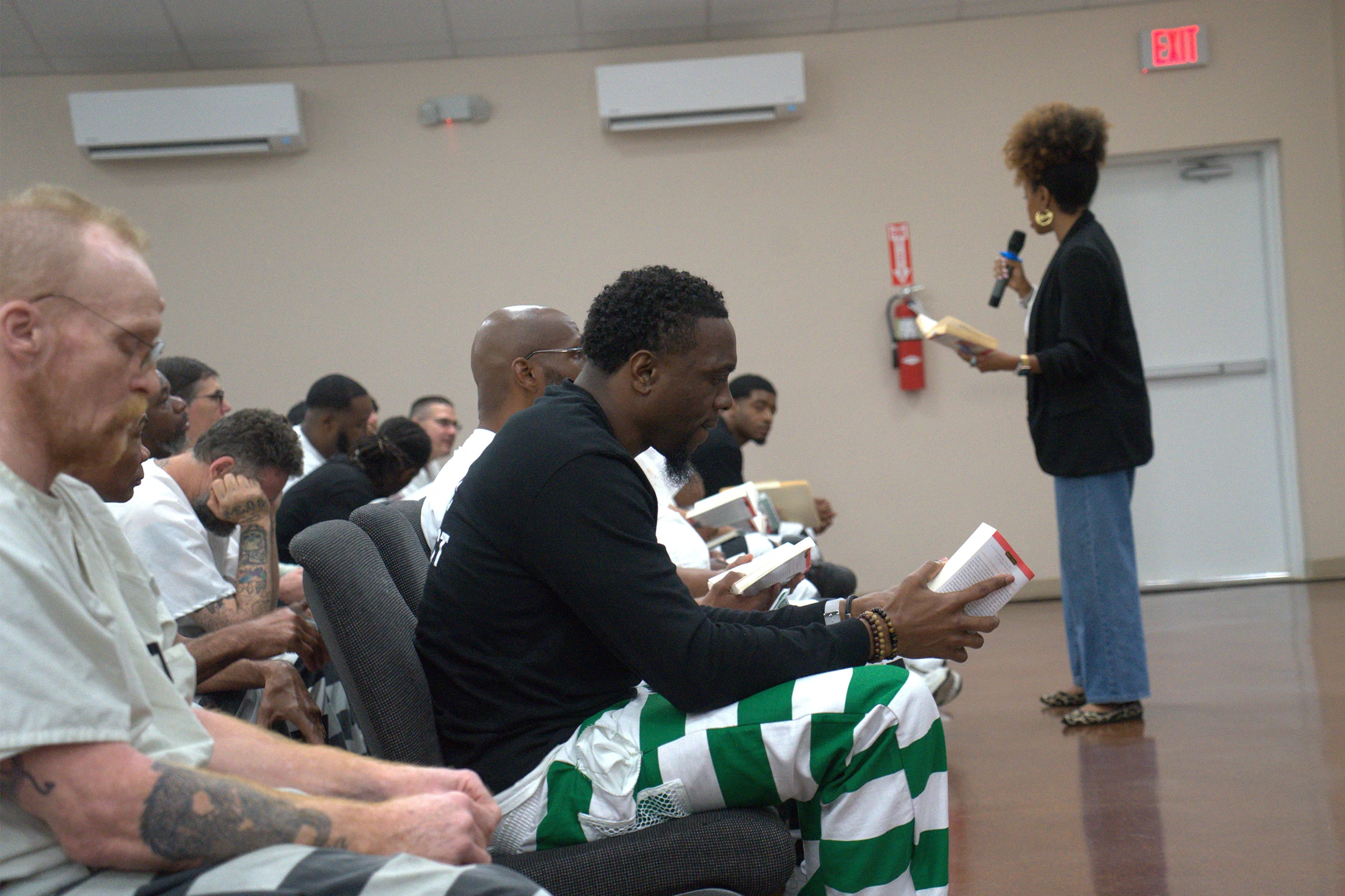 Unos hombres encarcelados se sientan a leer libros mientras una mujer dirige una discusión al frente de una habitación luminosa con paredes beige y una puerta de salida de incendios.