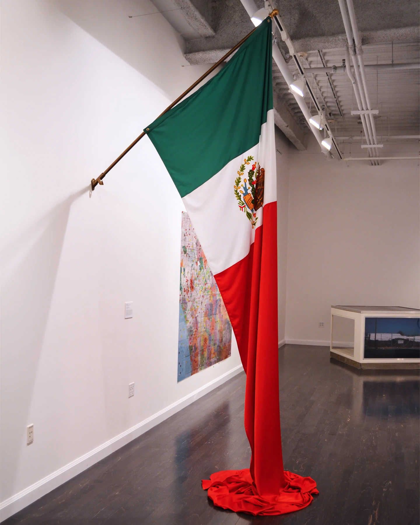 An extra long Mexican flag hanging from a holder on a white wall inside of a gallery space