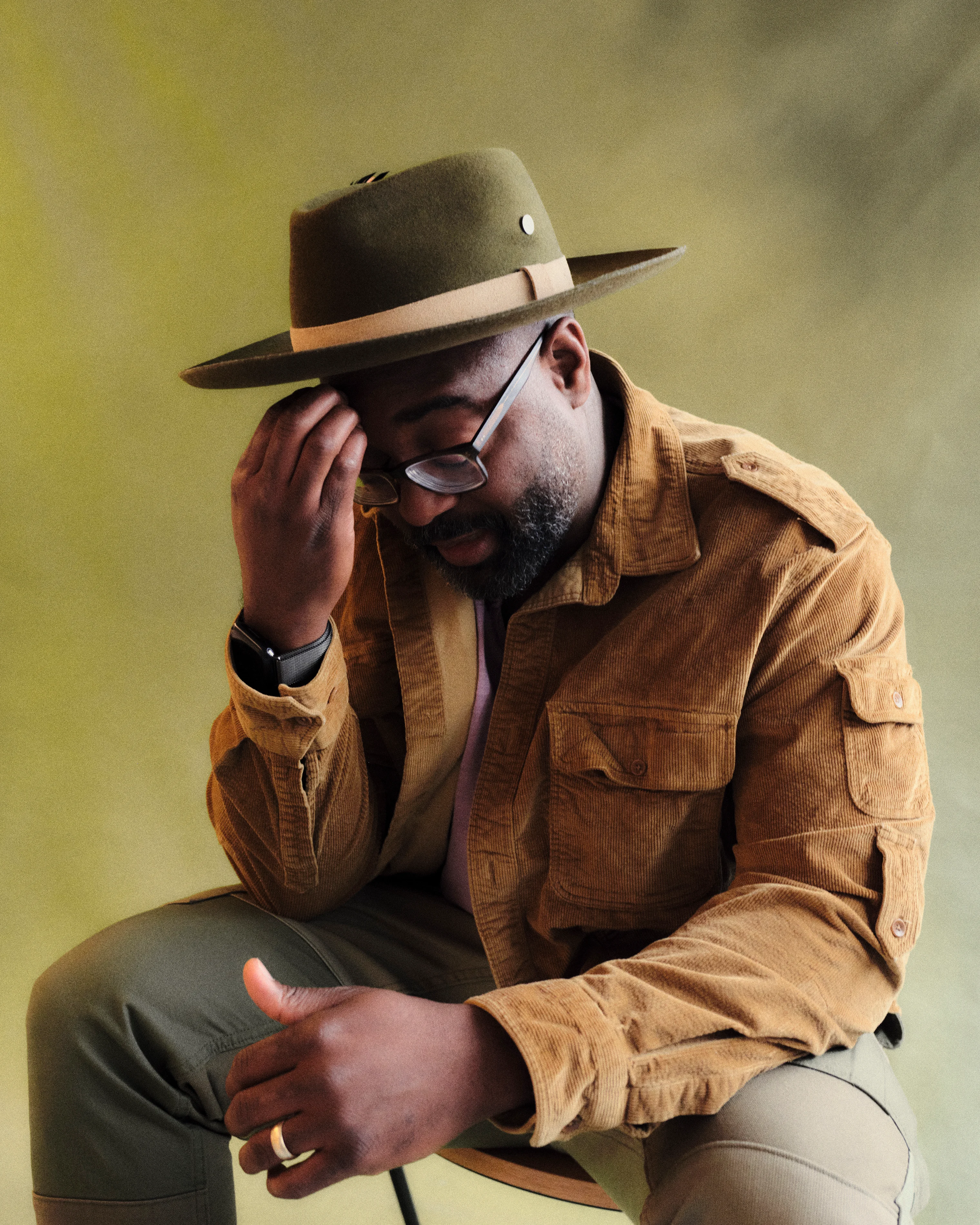 A man in a brown jacket and green hat sits on a stool against a green background, touching his glasses with one hand and resting the other on his lap.
