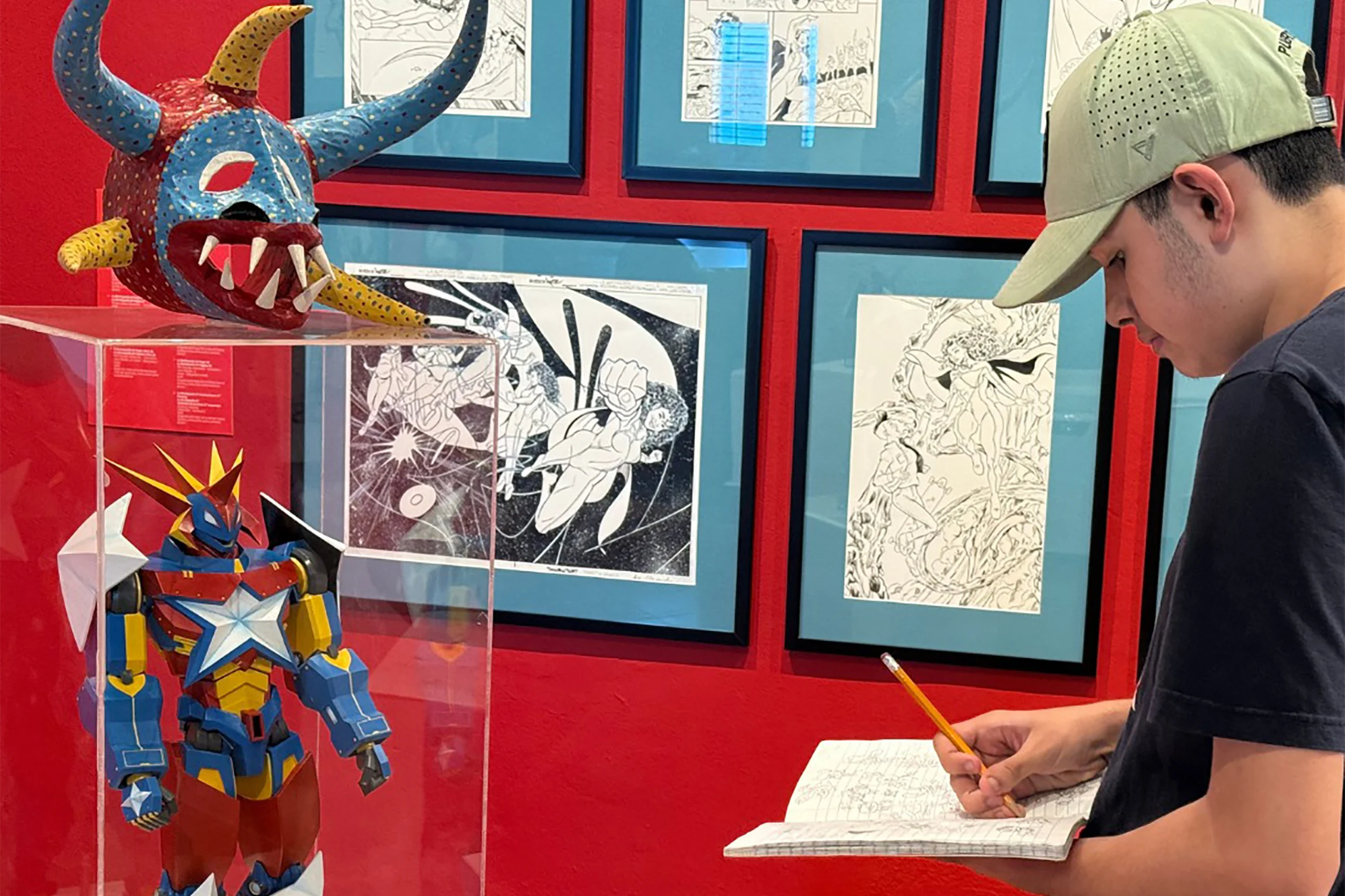 Un adolescente dibuja cerca de una máscara colorida, una escultura de robot y un cómic enmarcado exhibido en una pared de un rojo intenso.