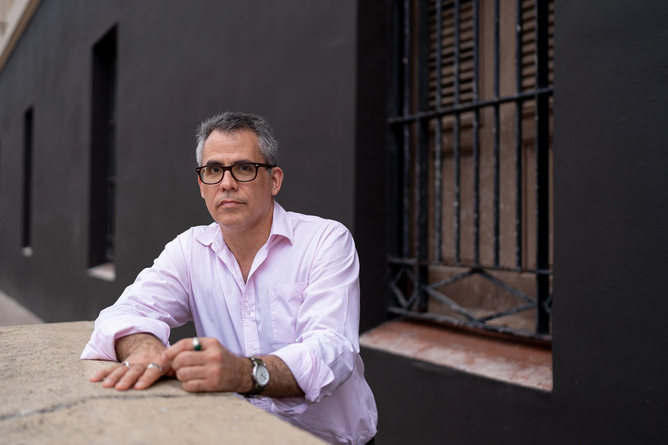 Francisco Font Acevedo wearing a pink dress shirt and glasses standing outdoors in front of a black painted wall leaning on a low concrete barrier