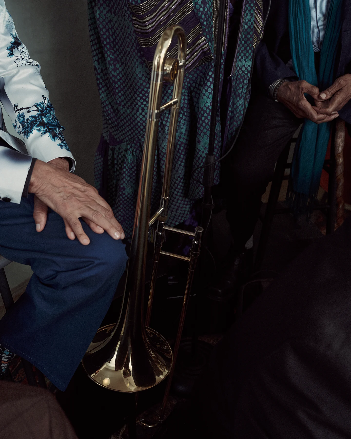 Close-up of a brass trombone standing upright between seated musicians. Hands and colorful clothing frame the instrument, with a microphone stand and dark studio floor visible in the background.