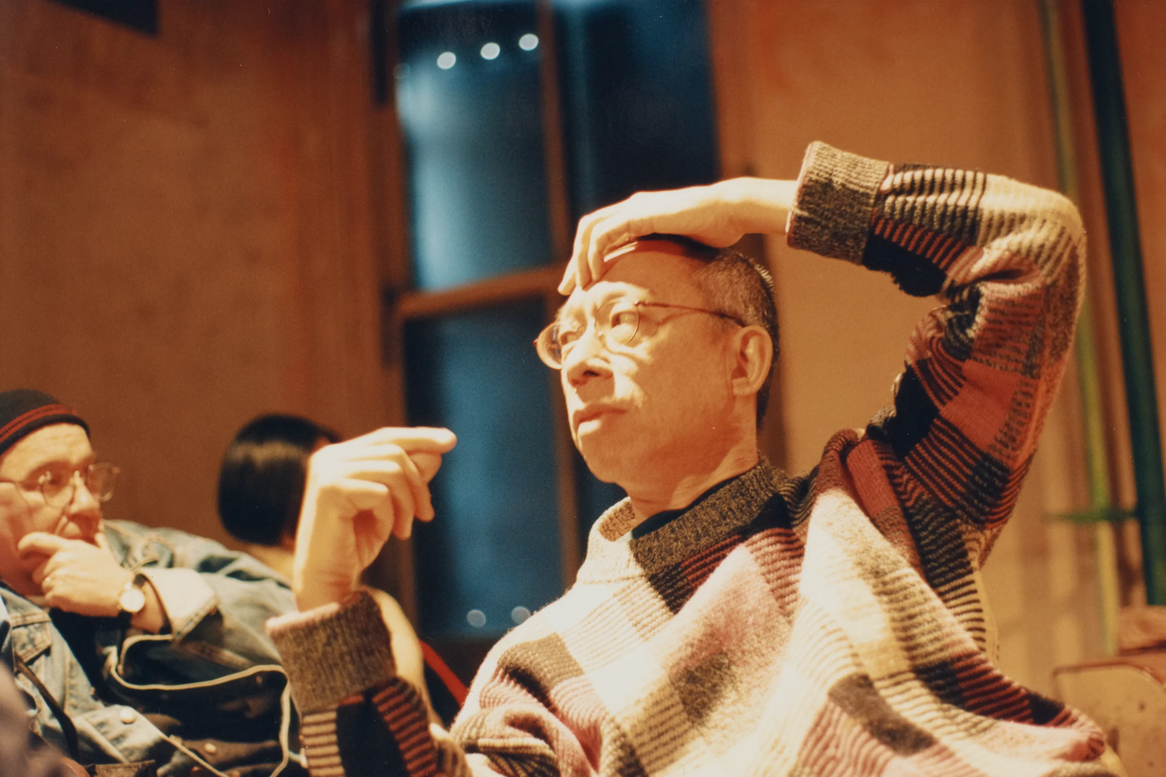 A man wearing glasses and a patterned sweater sits indoors, touching his forehead thoughtfully, with two other people visible in the background.