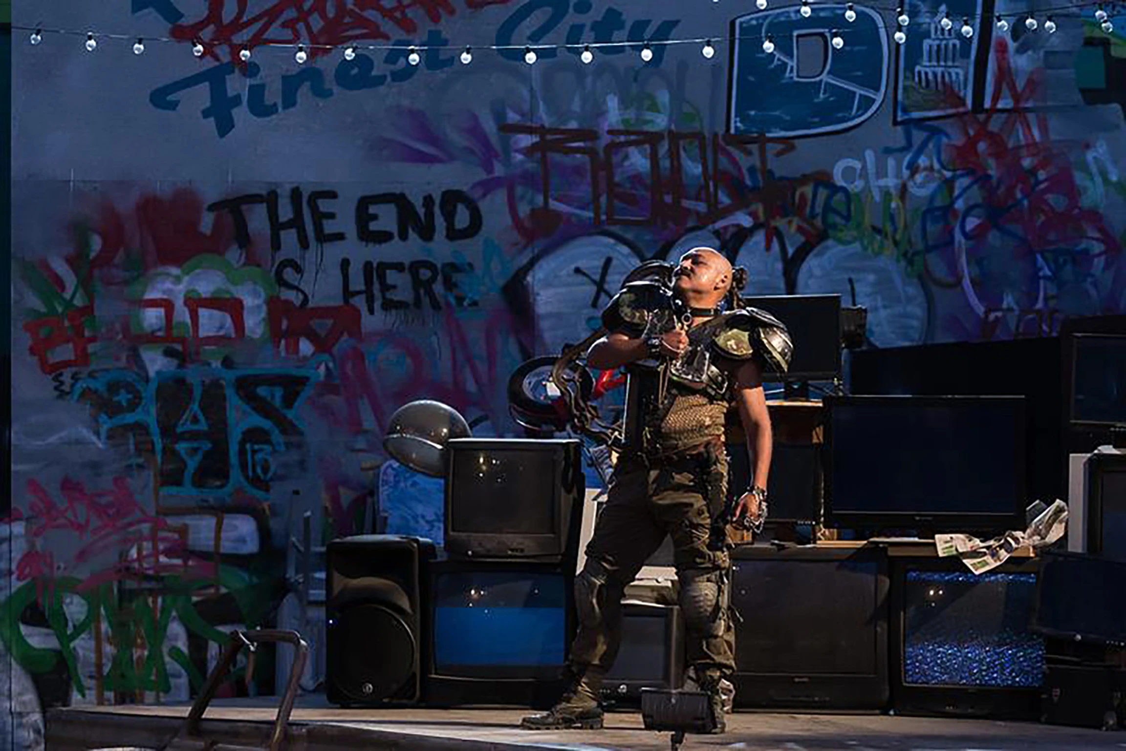A performer on a stage dressed in urban clothes and football pads in front of a wall covered in graffiti 