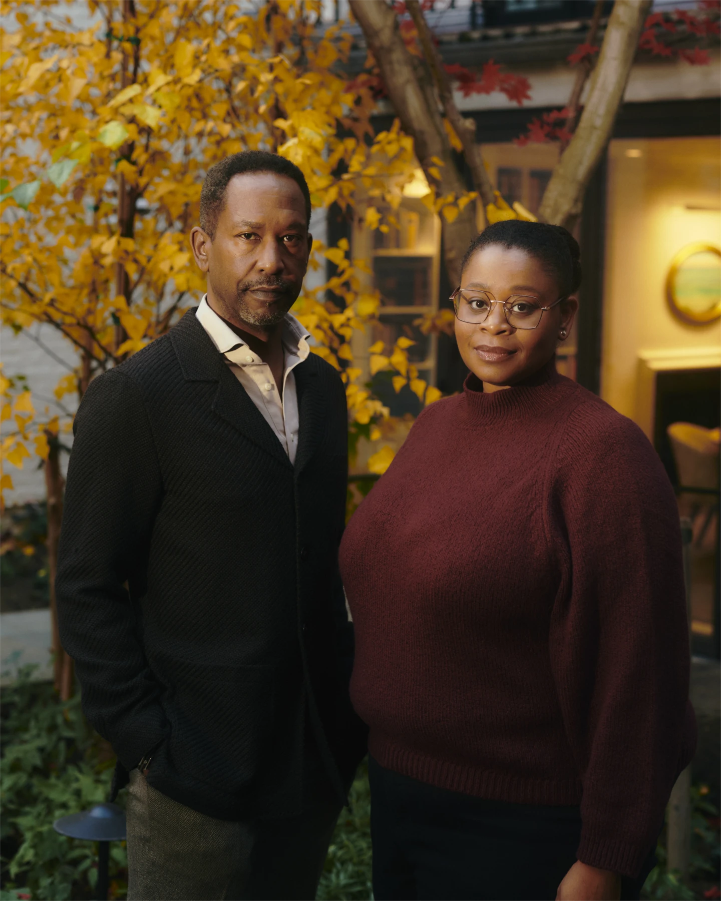 An African American man in a dark sport coat stands on the viewers left, and on the right is an African American woman in a maroon sweater with glasses. They are in front of a gold leafed tree.