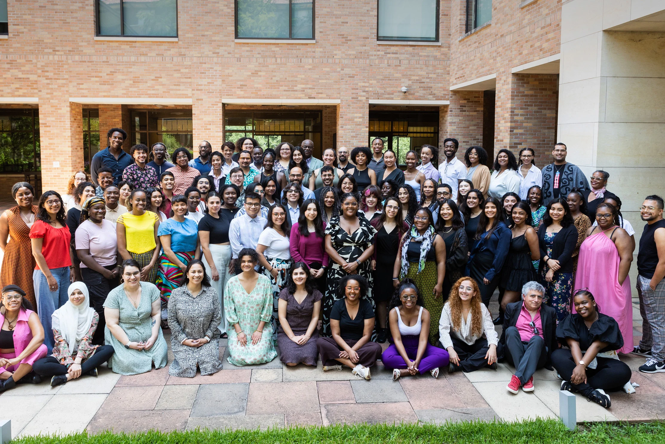 Group photo of about 80 diverse individuals smiling and posing in rows in an outdoor courtyard with brick walls and large windows. The group is dressed in casual and semi-formal attire, suggesting a celebratory or professional event.
