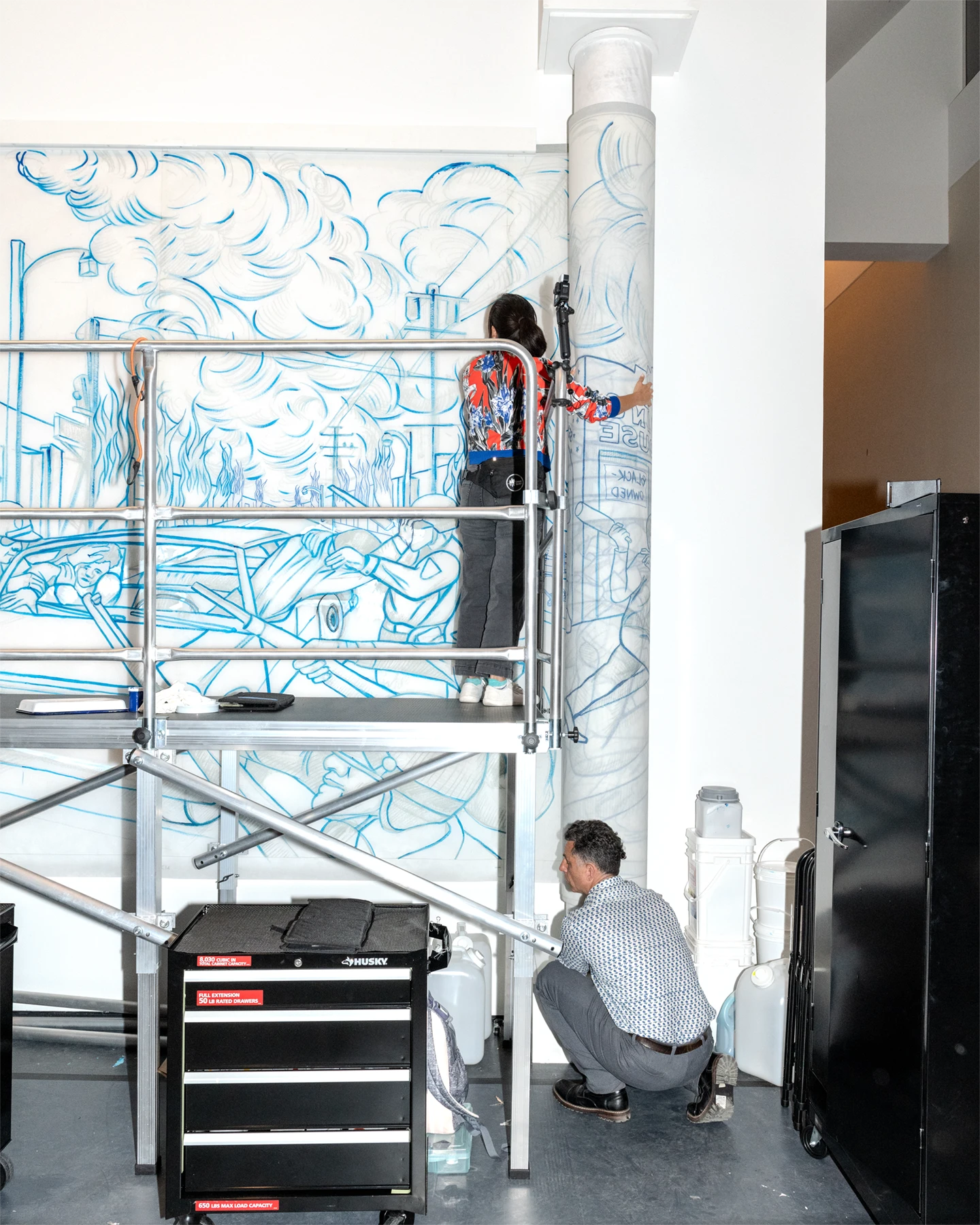 An artist standing on scaffolding while installing a sketched artwork on a wall with another person crouches on the floor next to it