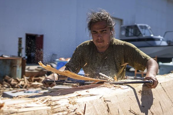 An Indigenous carver is using a tool to shave wood from a large piece of wood that will be shaped to be a totem pole.