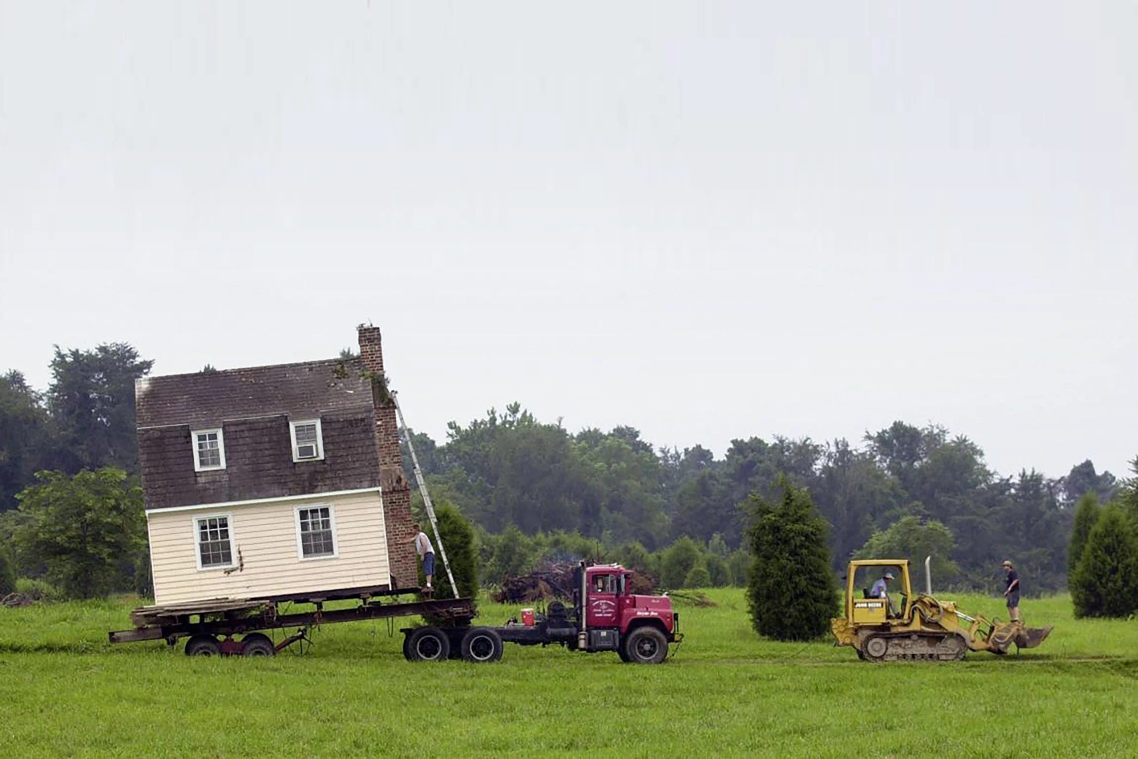 In an open field, a truck tows a fully constructed house on a flatbed.