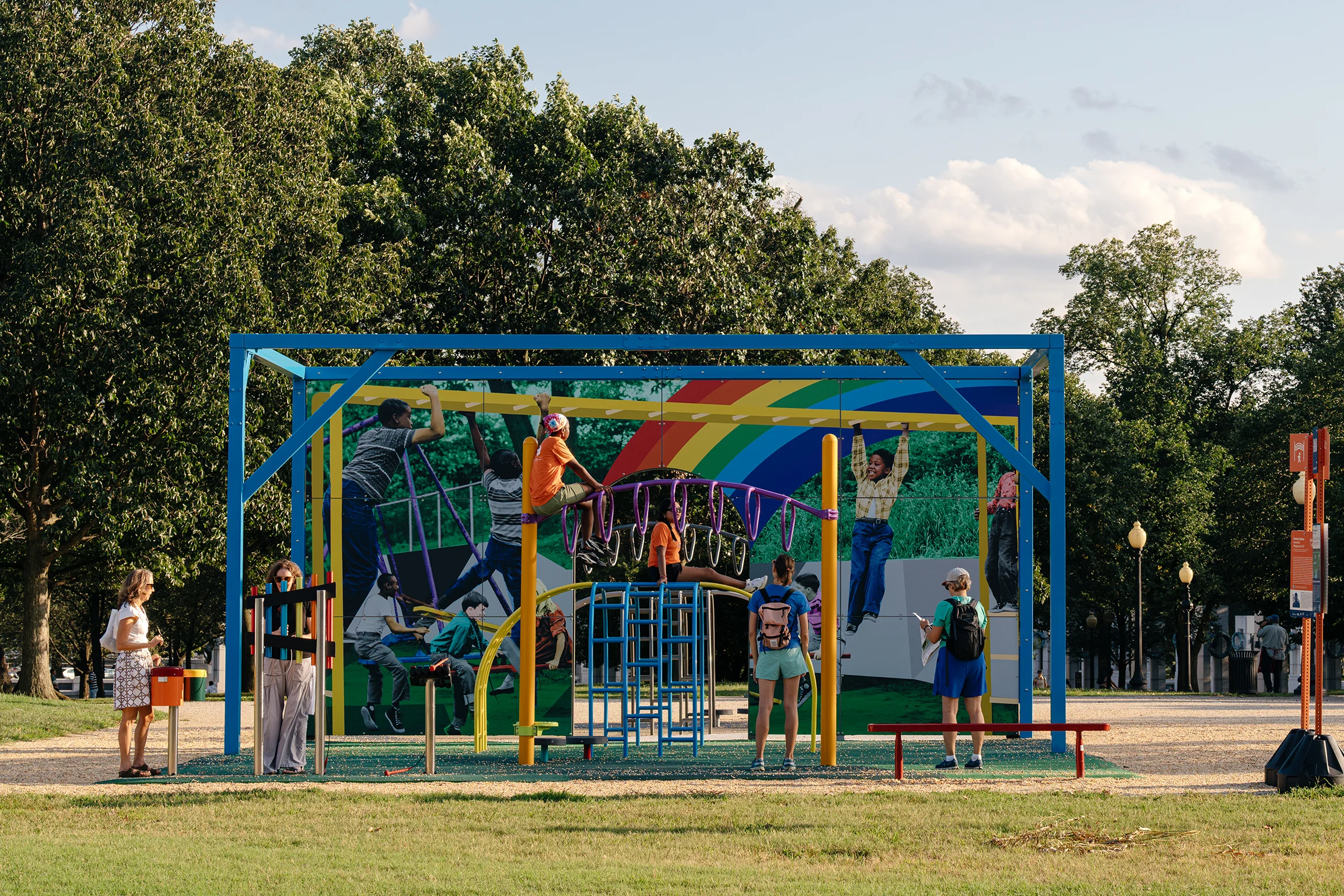 A colorful playground with many children playing on it in front of a colorful mural with a rainbow painted on it inside of a park lined with trees.
