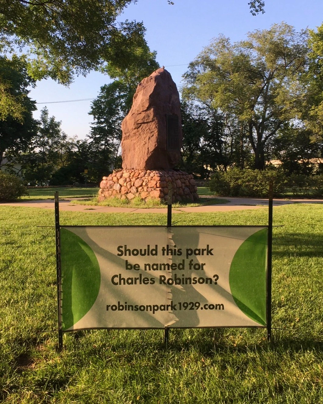 In a clearing, a tall red boulder sits on a pedestal. The boulder is almost as tall as the trees around it. Posted in the ground in front of the boulder, a sign reads "Should this park be named for Charles Robinson? robinsonpark1929.com"