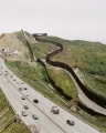 In portrait orientation, an aerial view of a major highway running parallel to a large border wall set on a hilly landscape