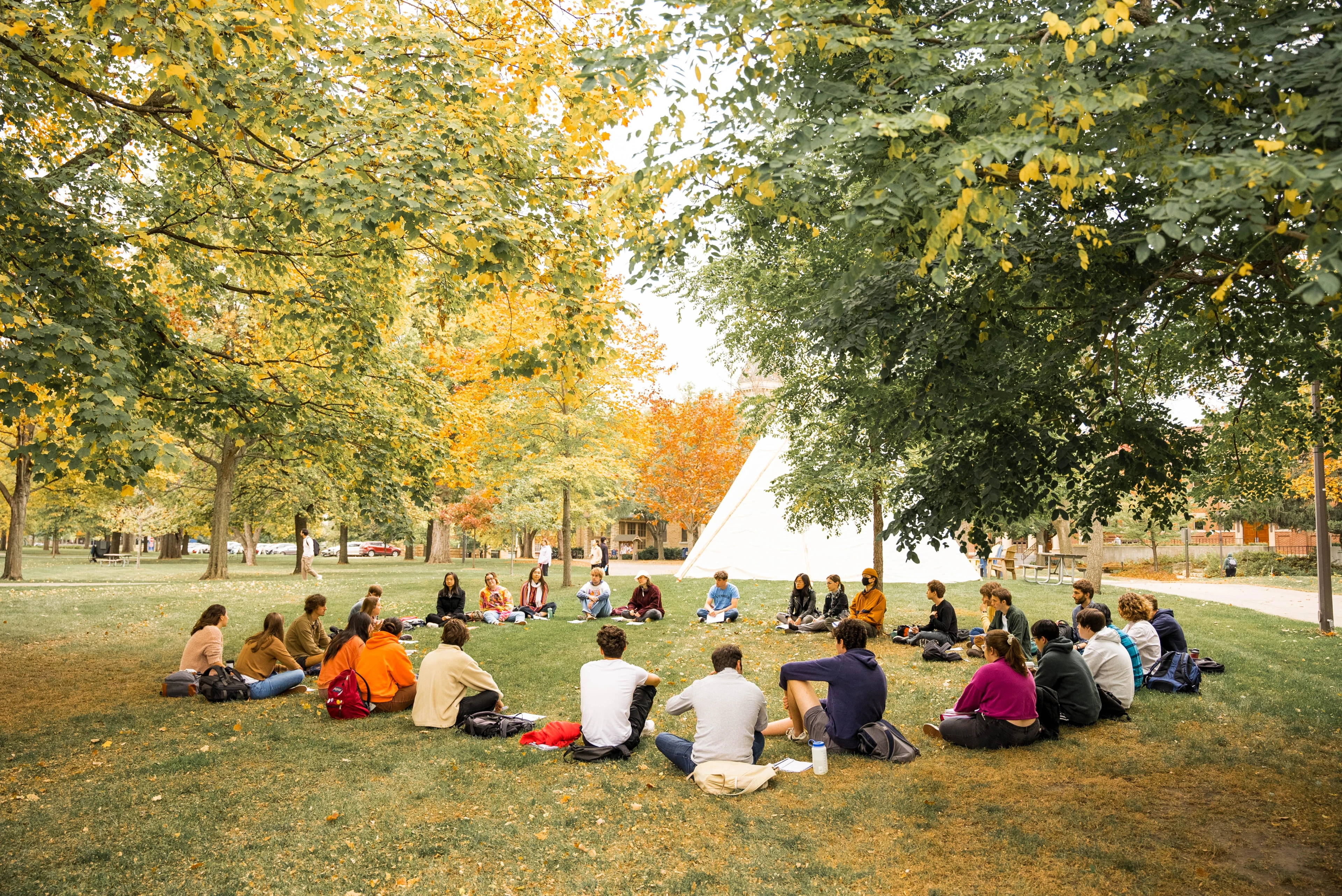 A group of students sits in a circle in the grass underneath trees on a sunny day