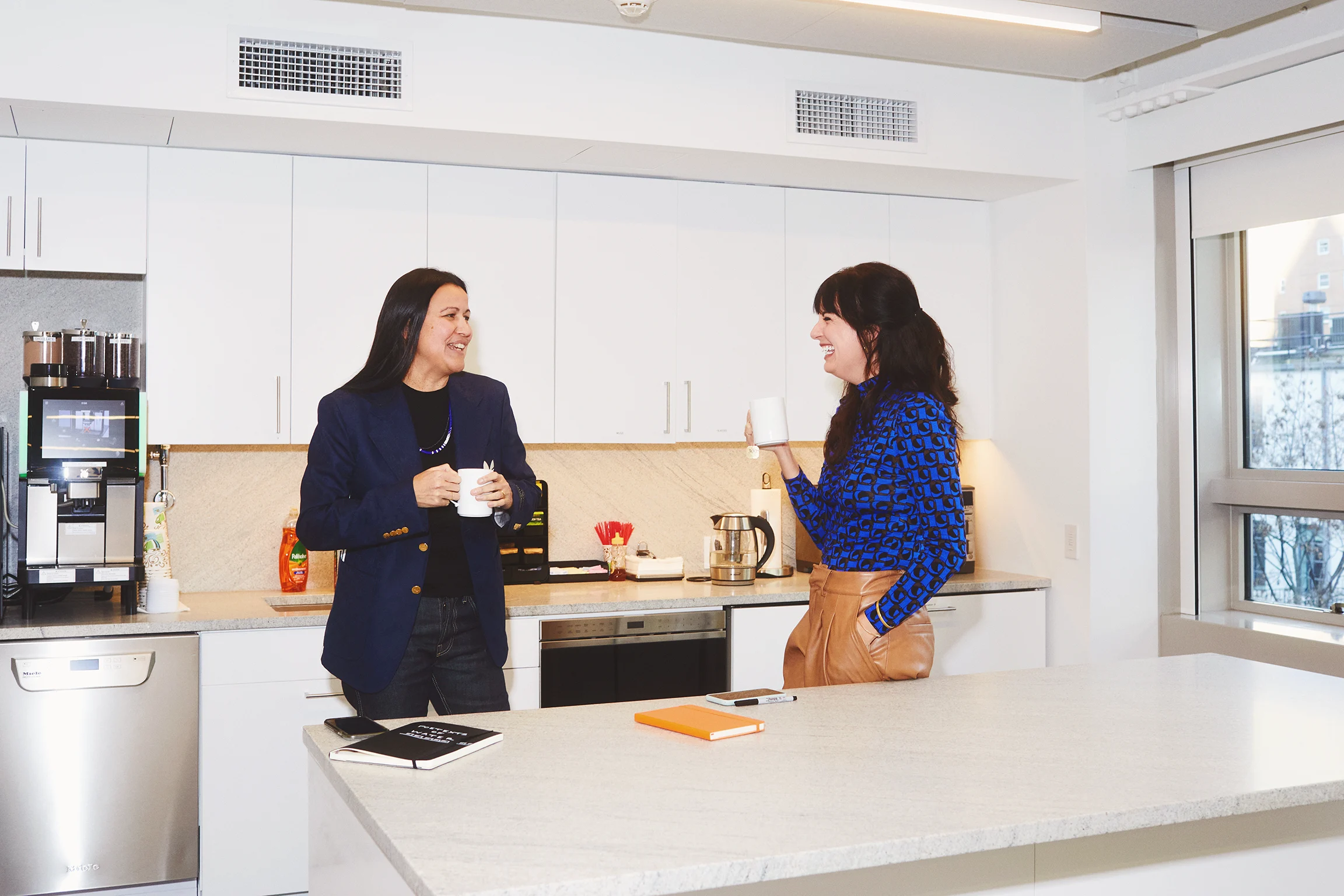 An Indigenous scholar holds a mug while standing in a kitchenette in conversation with a grantmaker.