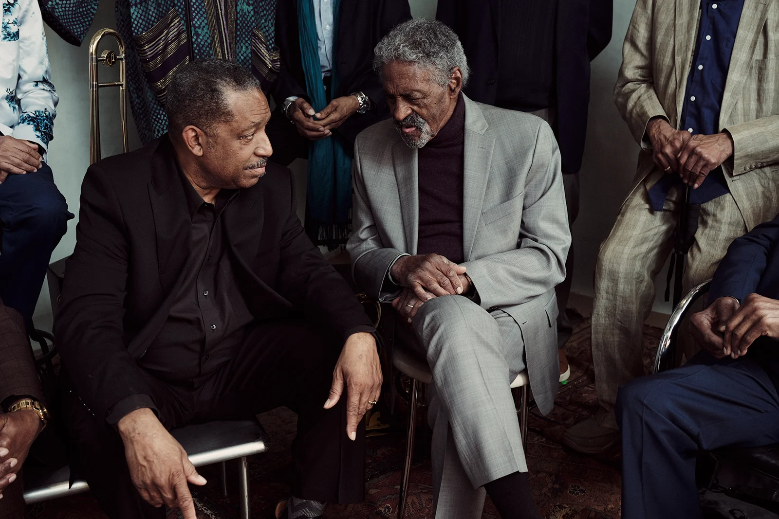 Two older musicians in suits sit facing each other in conversation, surrounded by fellow band members. A trombone stands beside them, and the close composition highlights their interaction in a studio setting.