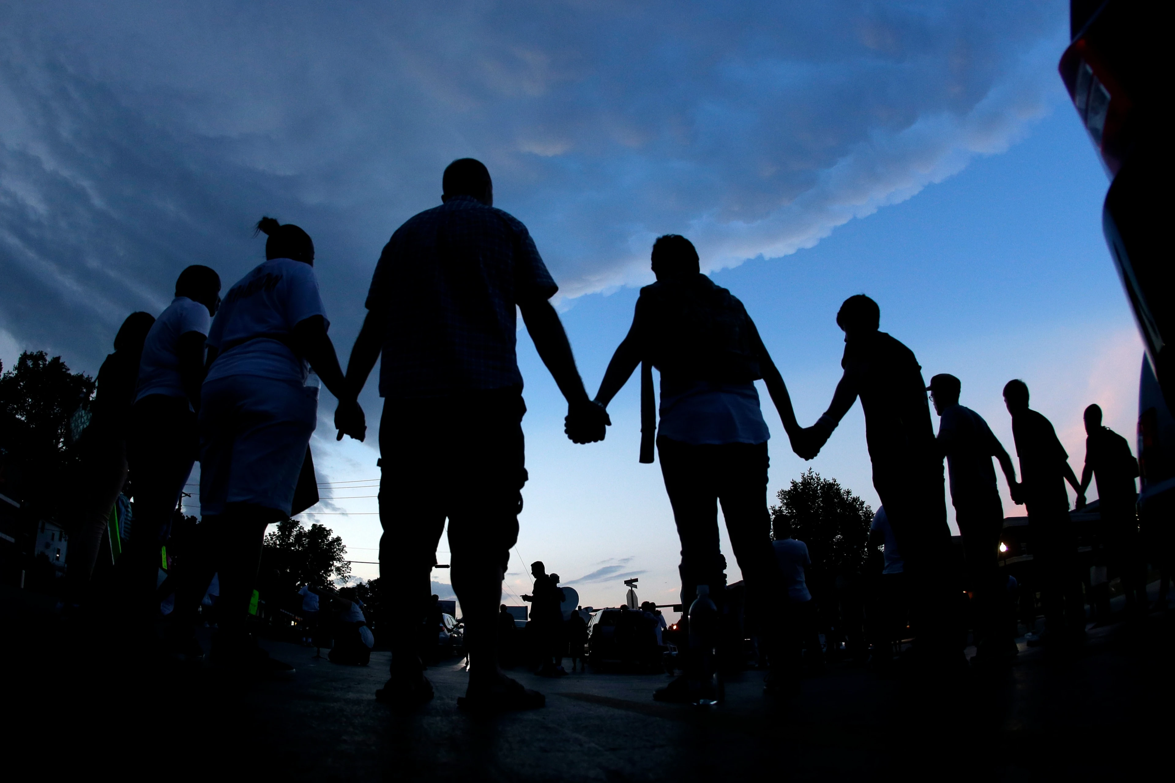 Silhouettes of people standing in a dusk sky. People are holding hands in a chain.