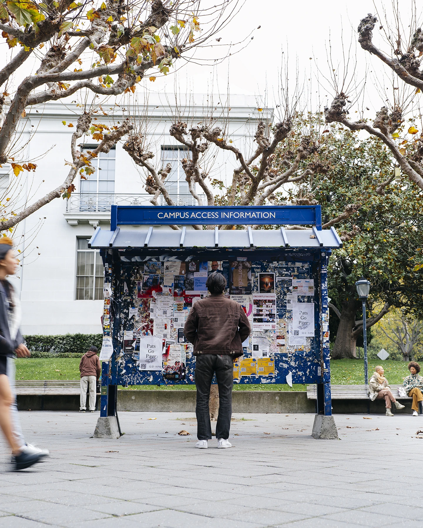 A man in a brown jacket stands in front of blue bulletin board that is covered with papers and advertisements  