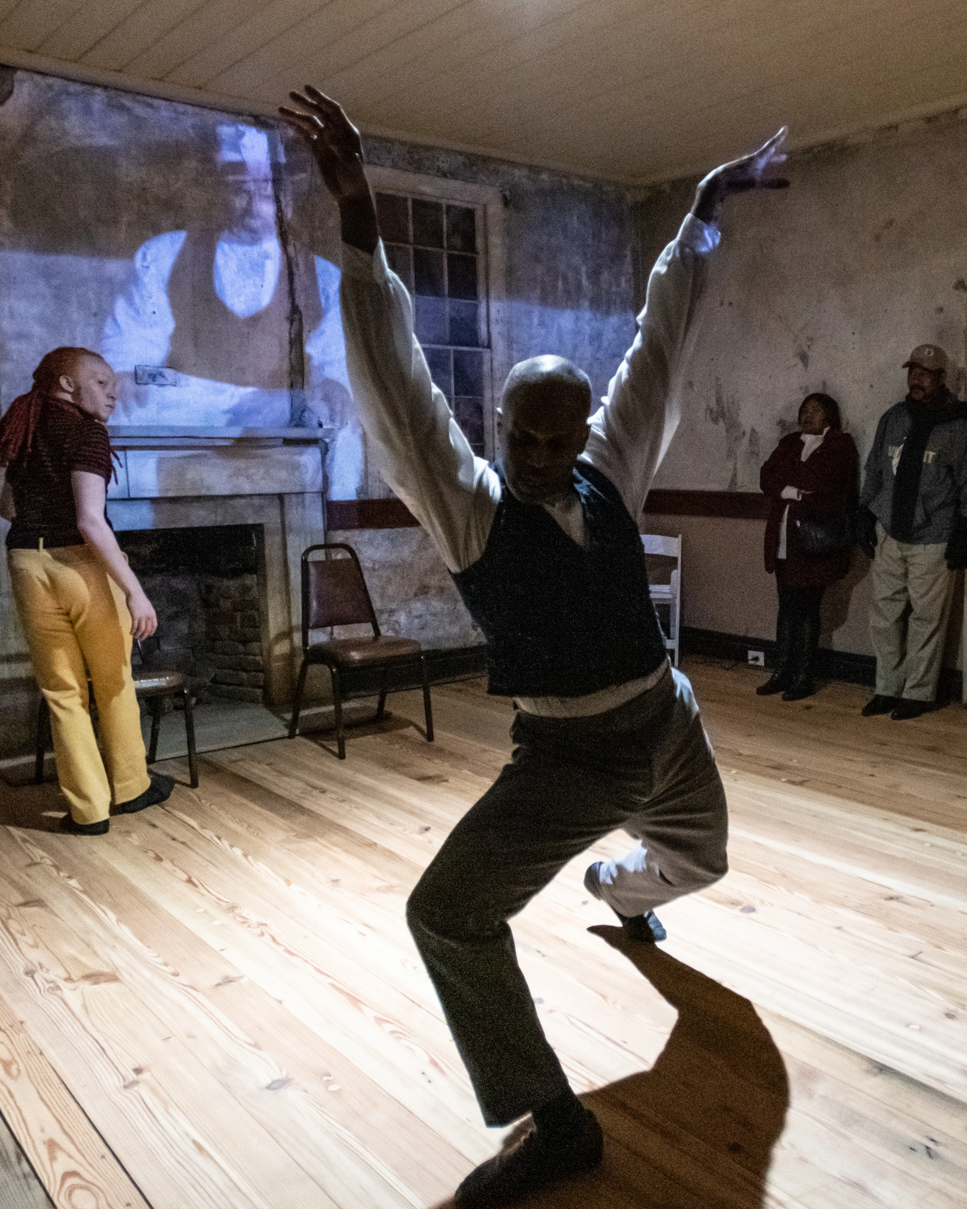  A single dancer performs in Wideman Davis Dance’s “Migratuse Ataraxia.” They are raising their arms and stepping their right foot forward. A multimedia projection is displayed behind them.
