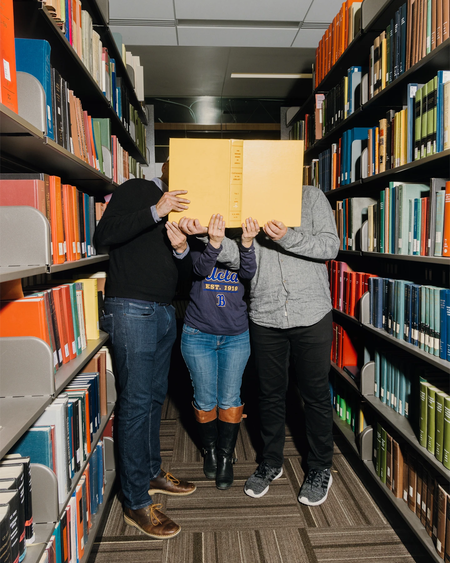 Three students in a college library stand in the stacks of books with a large book covering their faces