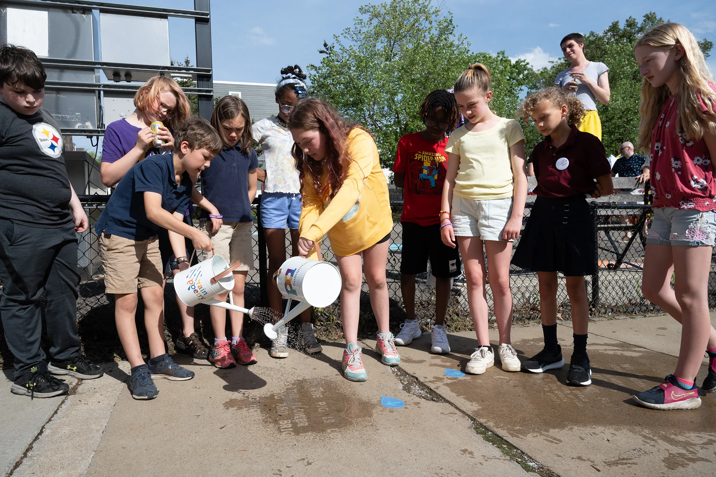 Los niños vierten agua en una acera para revelar poemas ocultos, rodeados de compañeros de clase y maestros en un día soleado.