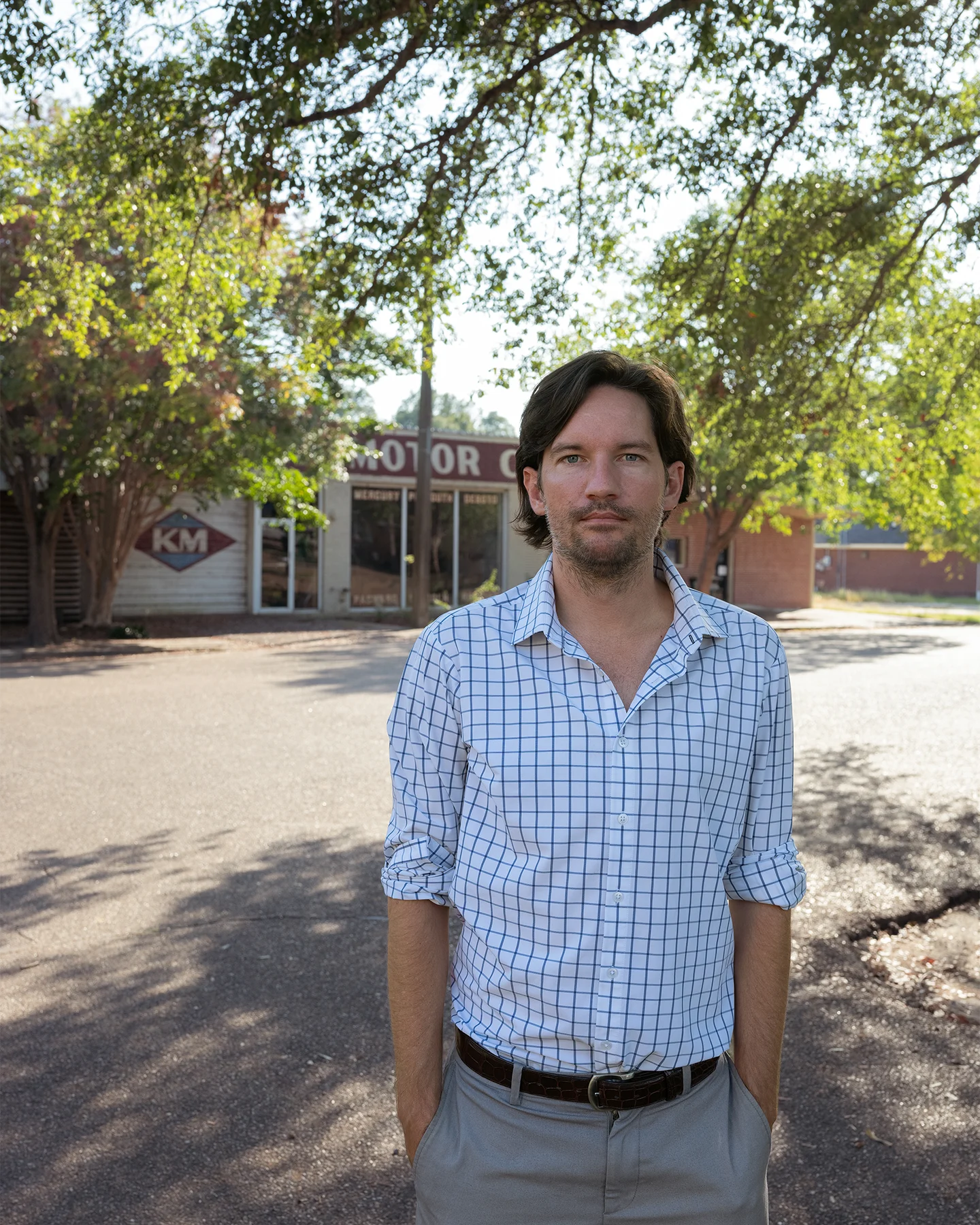 A white man with long dark hair dressed in a light blue checked shirt and gray pants standing with his hands in his pockets outside of a car repair shop