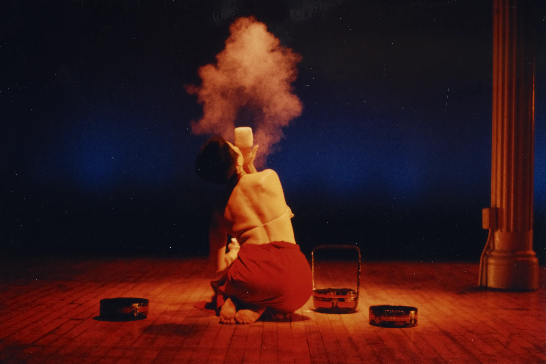 A person kneels on a wooden floor, holding a jar that emits a cloud of powder or smoke, surrounded by tambourines and lit by dramatic red lighting.