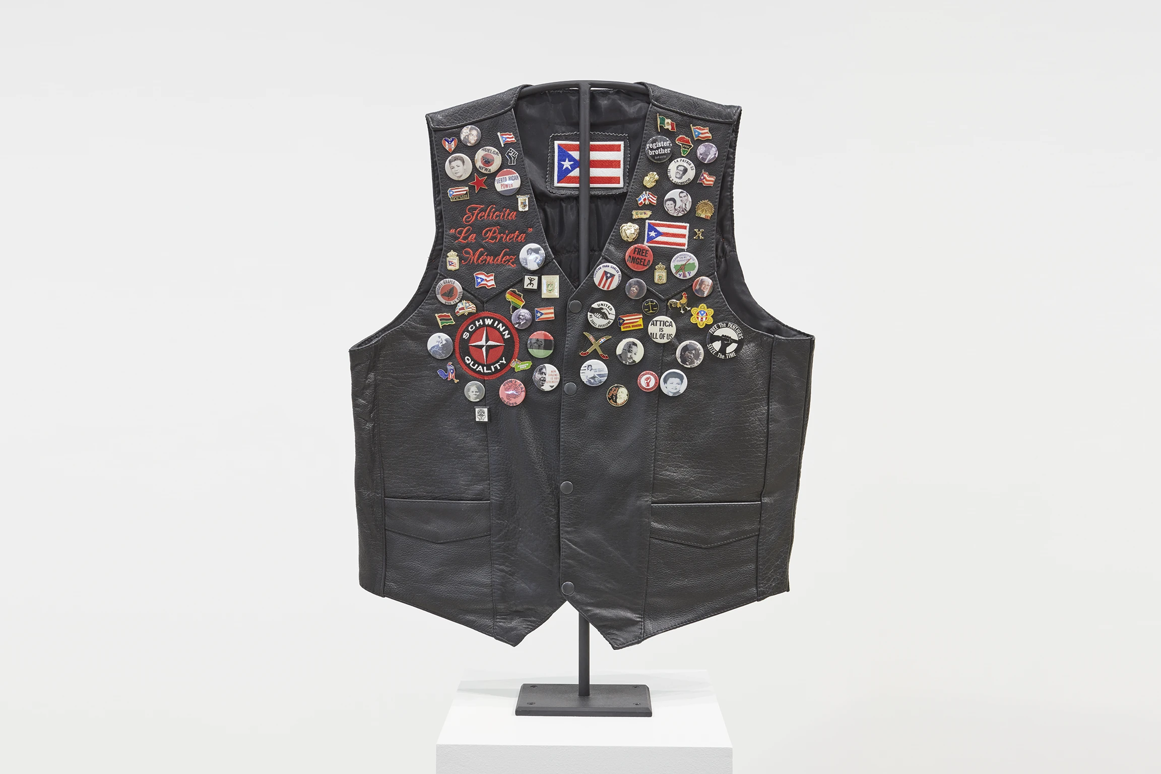 A black leather vest is adorned with colorful pins across the front of it. Embroidery on the vest reads: "Felícitas "La Prieta" Méndez" 