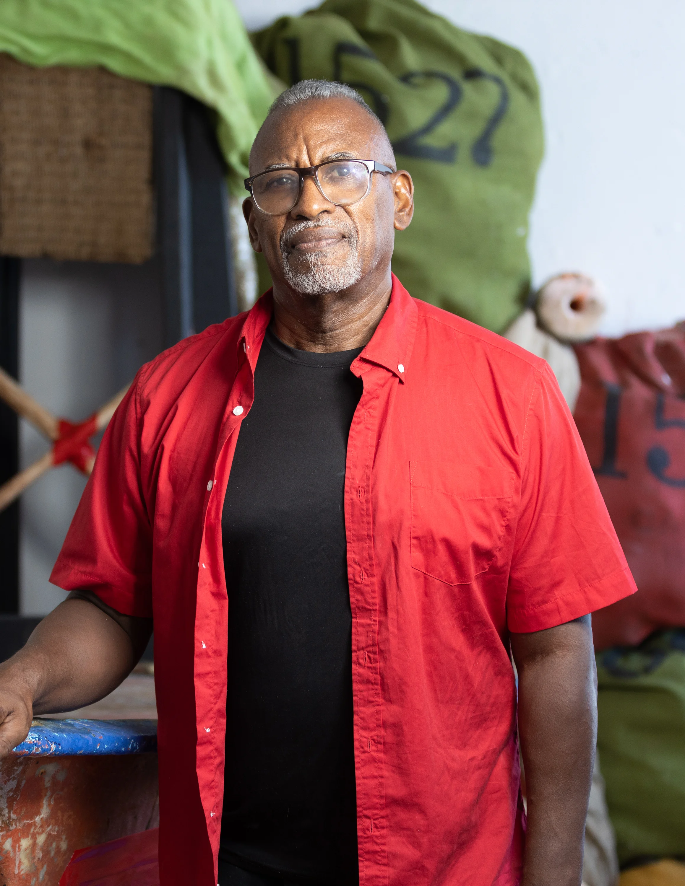 A male artist wearing glasses and a red button down shirt over a black tee shirt stands in front of an art installation