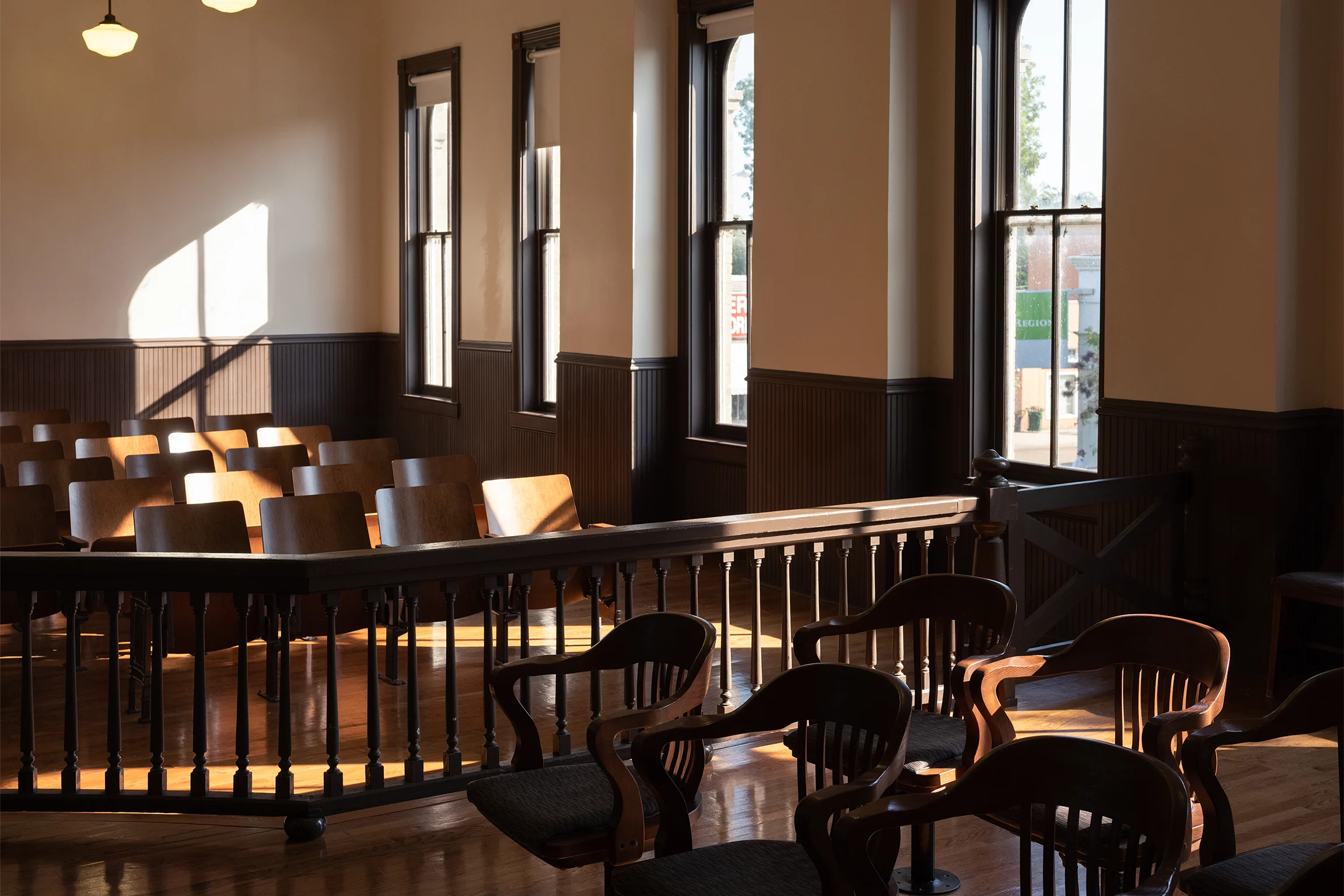 A view inside a courthouse room set with rows of wooden chairs and sunlight coming in from the tall windows on the right side of the room