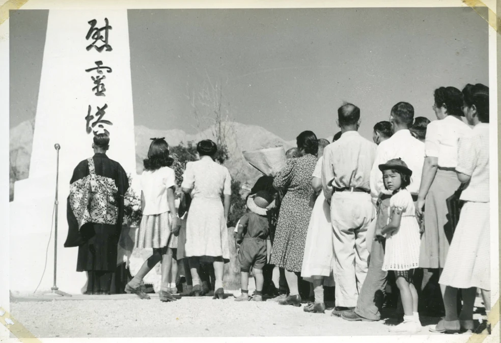 In black and white, a line of Asian individuals of all ages stand in front of a pillar. In big black letters, three Japanese Kanji characters read "Soul Consoling Tower." A young child looks back towards the camera.