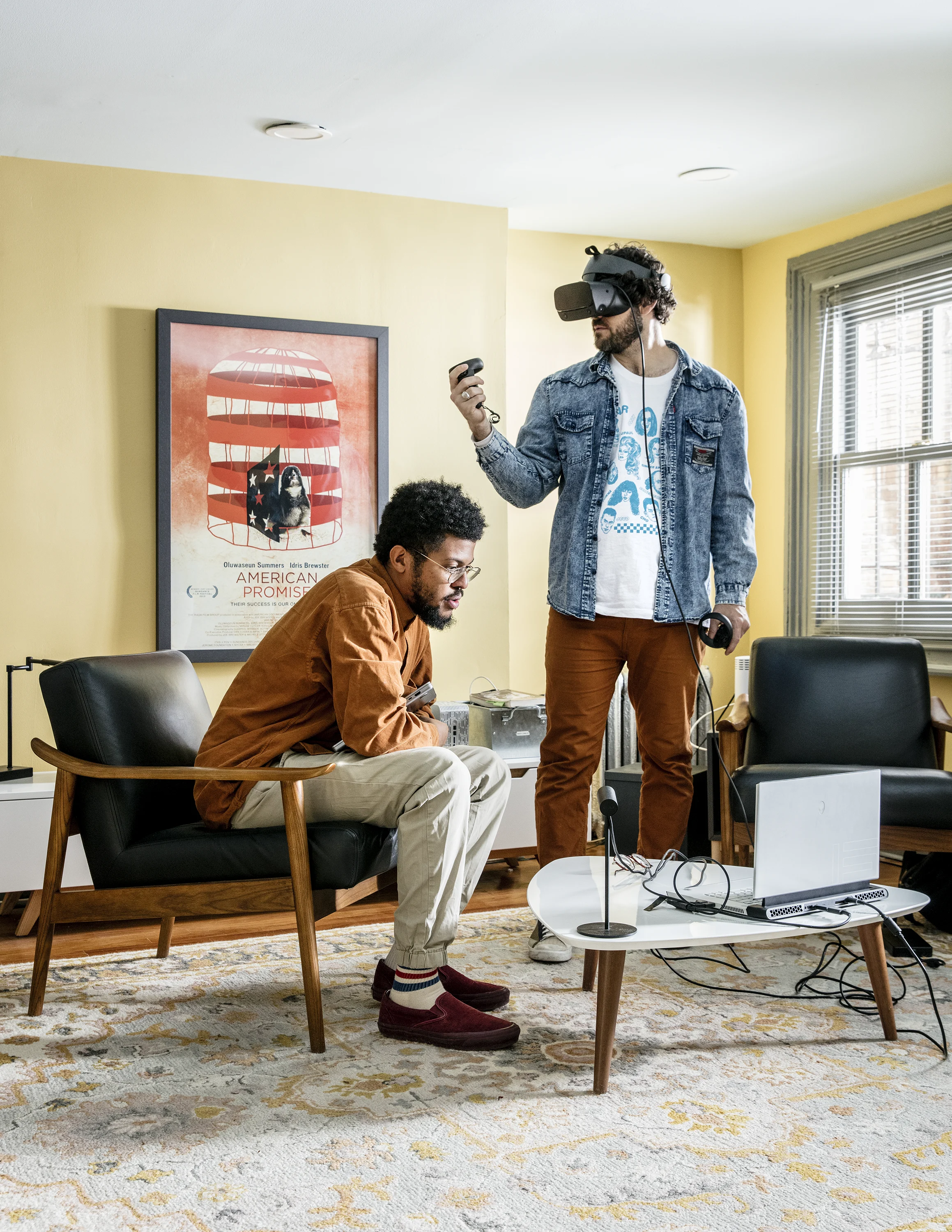 Two people, one sitting and one standing, in a living room are using a laptop and VR glasses 