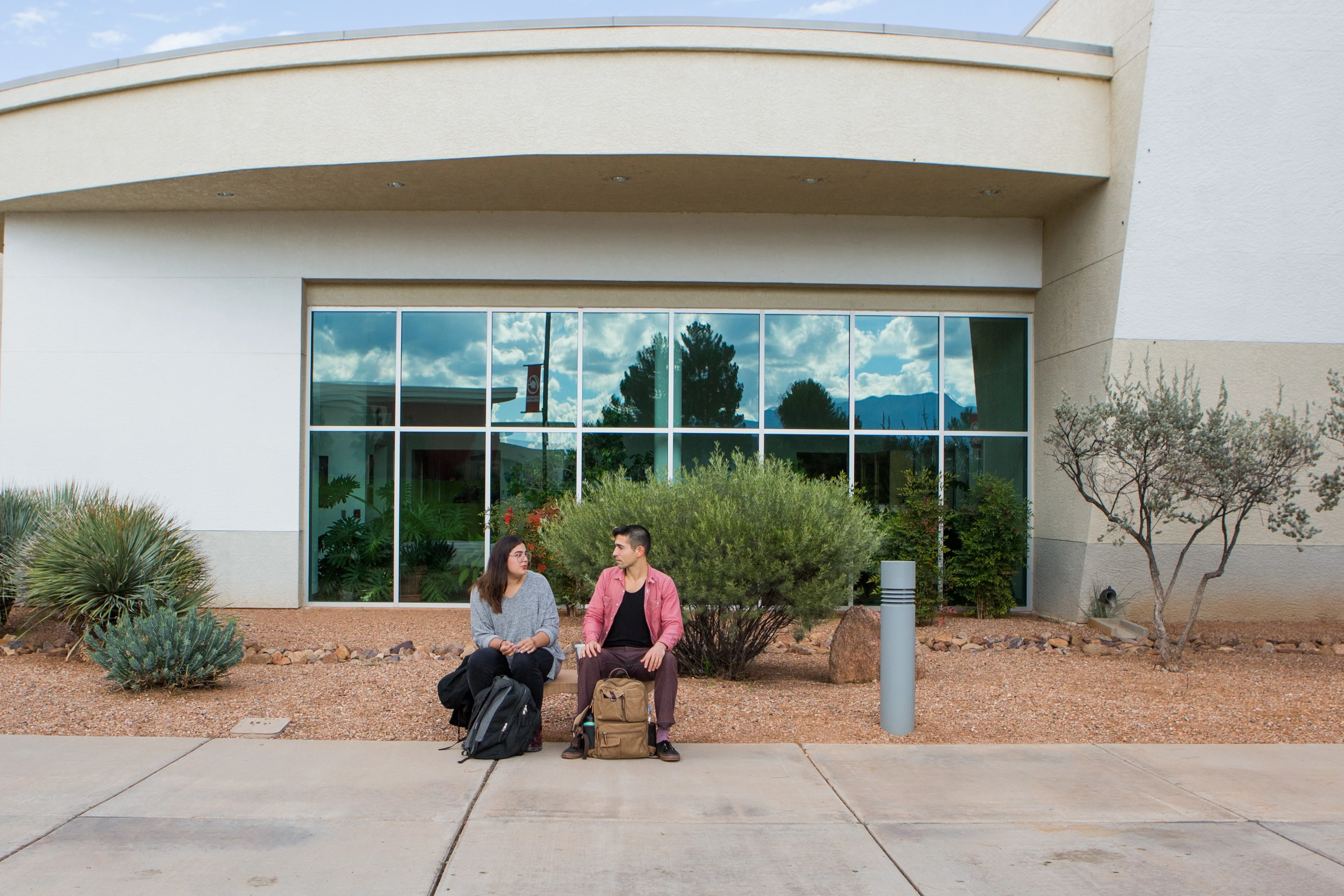 Two students sit in front of a building that is in the style of modern architecture. 