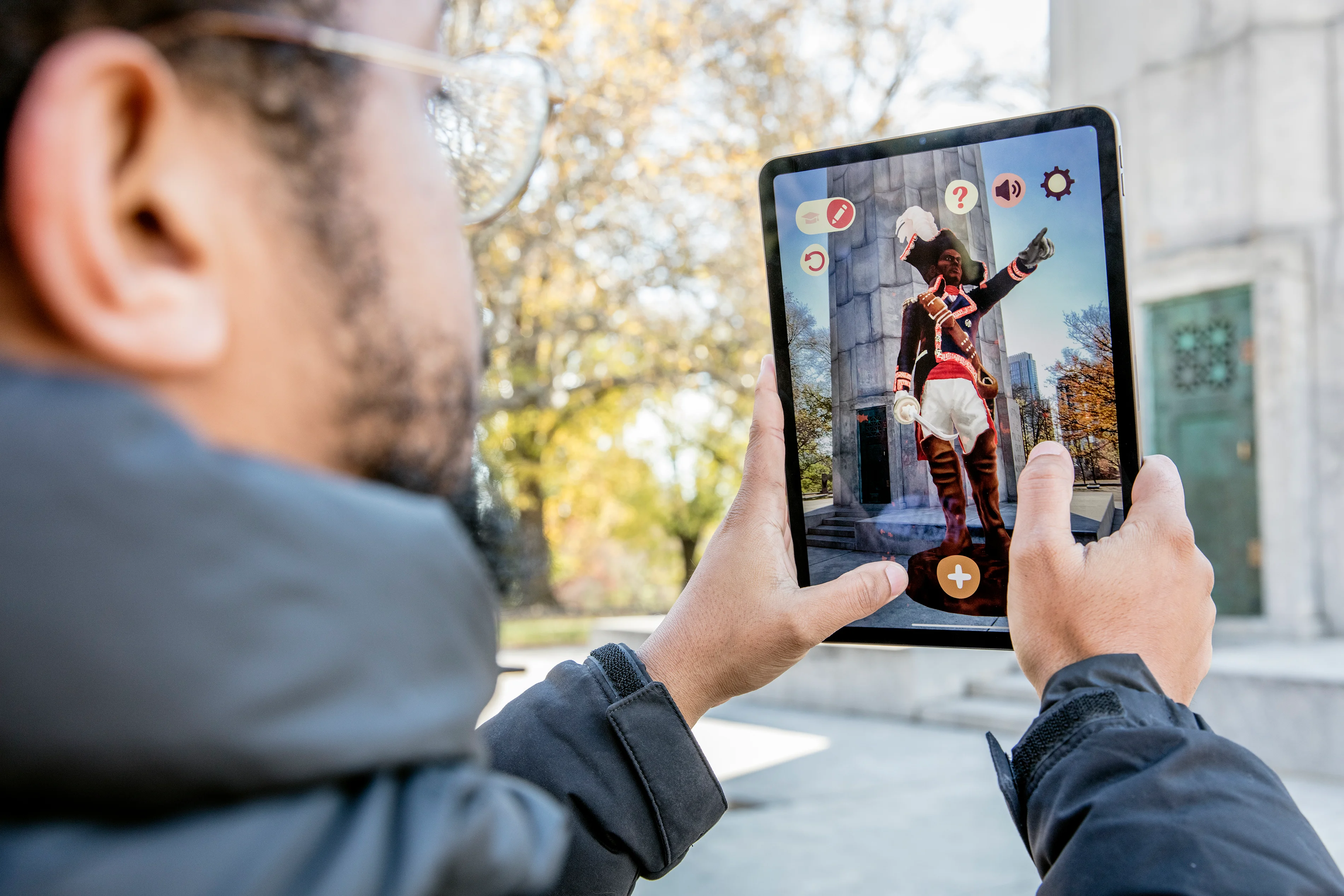 A man wearing glasses with a beard holds up an iPad to with an image of a colonial dressed figure standing in front of a concrete monument