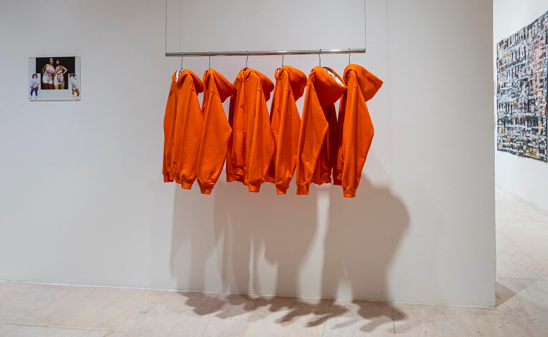 Six orange hooded sweatshirts hand in a gallery on display with other artworks