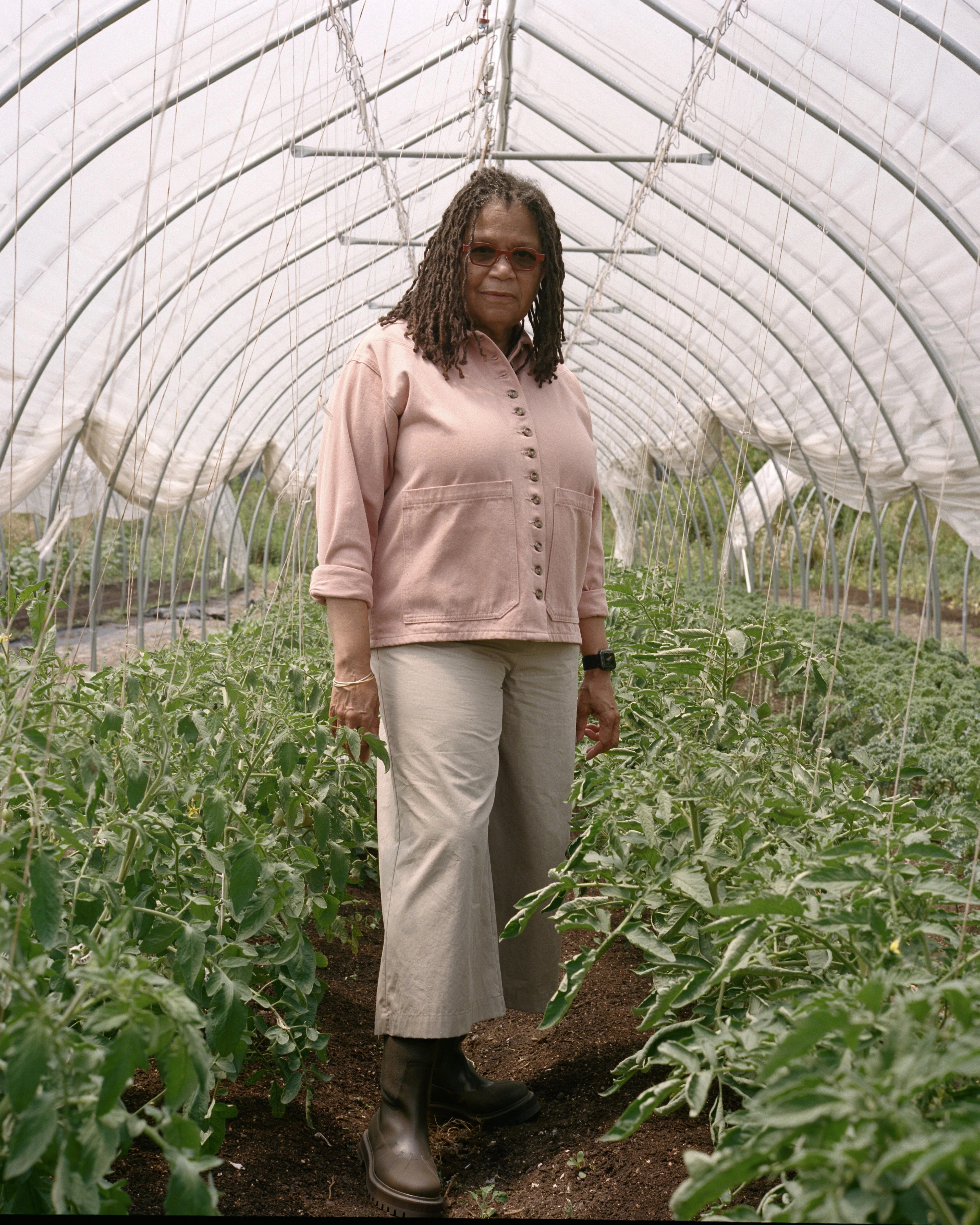 A Black woman stands in an indoor garden in a row of plants. She wears glasses and boots, looking at the camera.