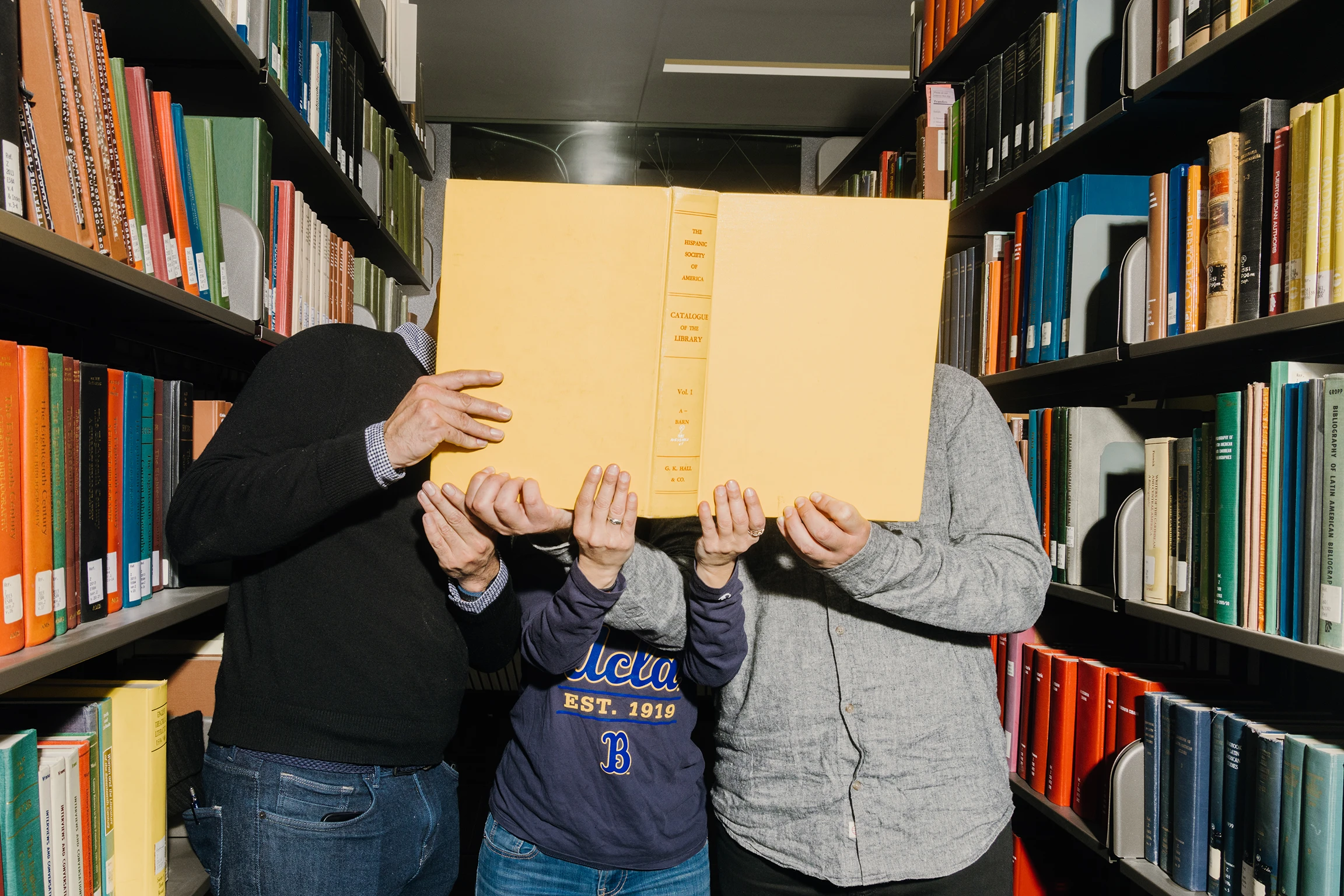 Three people stand in a library with shelves on either side of the group. Together, they hold a book in front of their faces so they are not visible to the camera.
