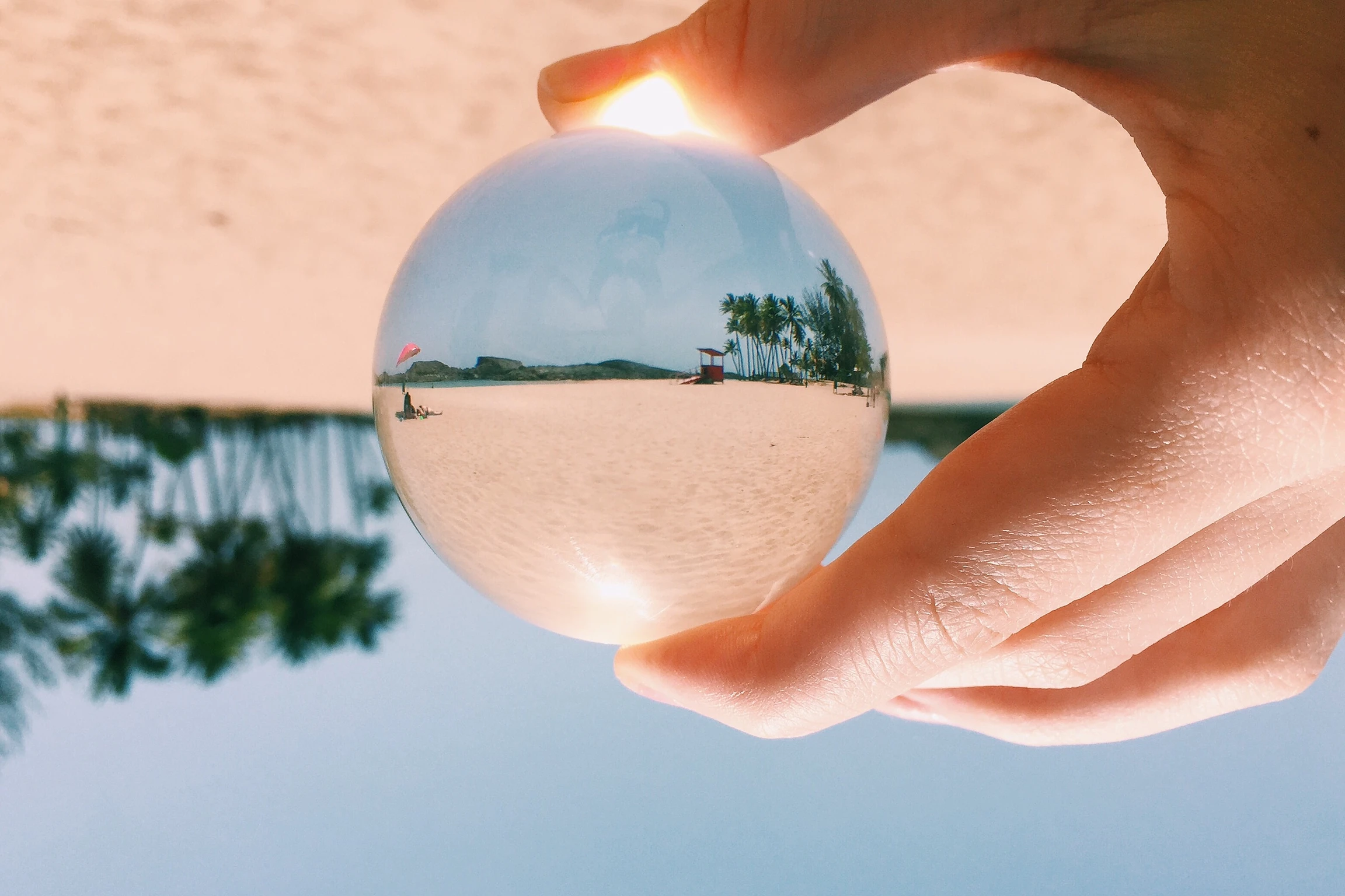 En primer plano, una mano sostiene un prisma a través del cual se refleja una escena de playa.