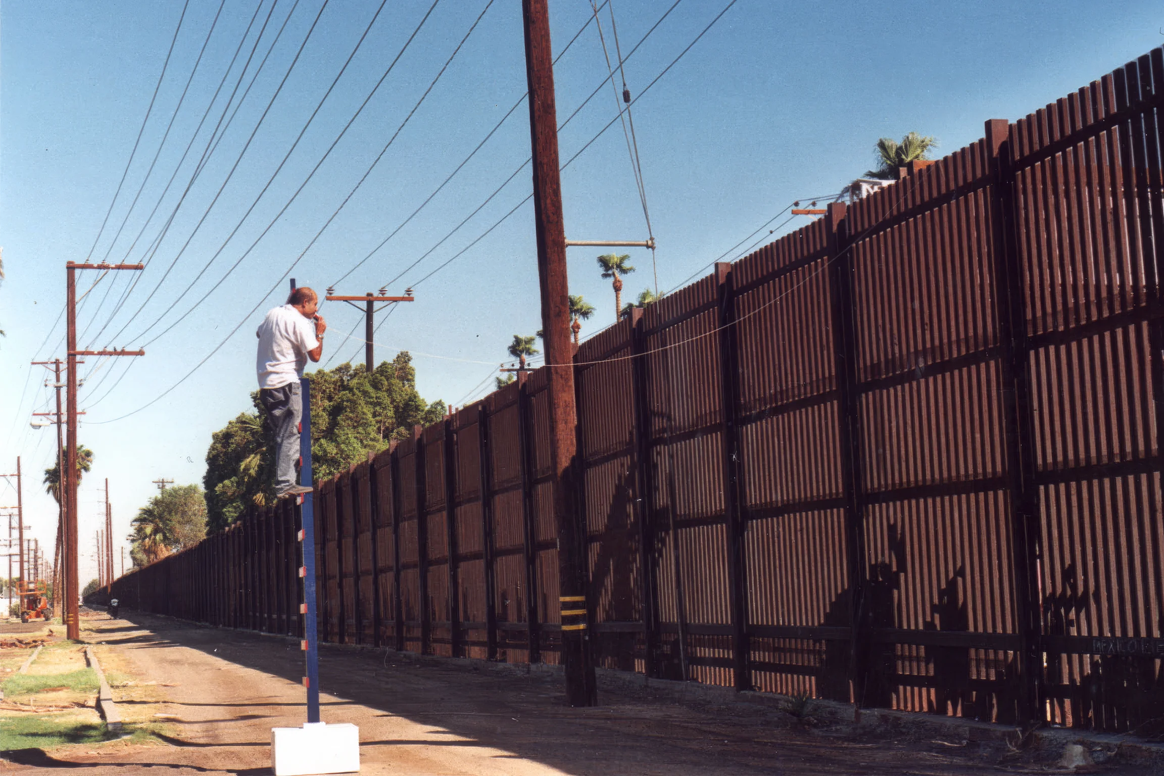 A man stands on a ladder overlooking a metal border wall