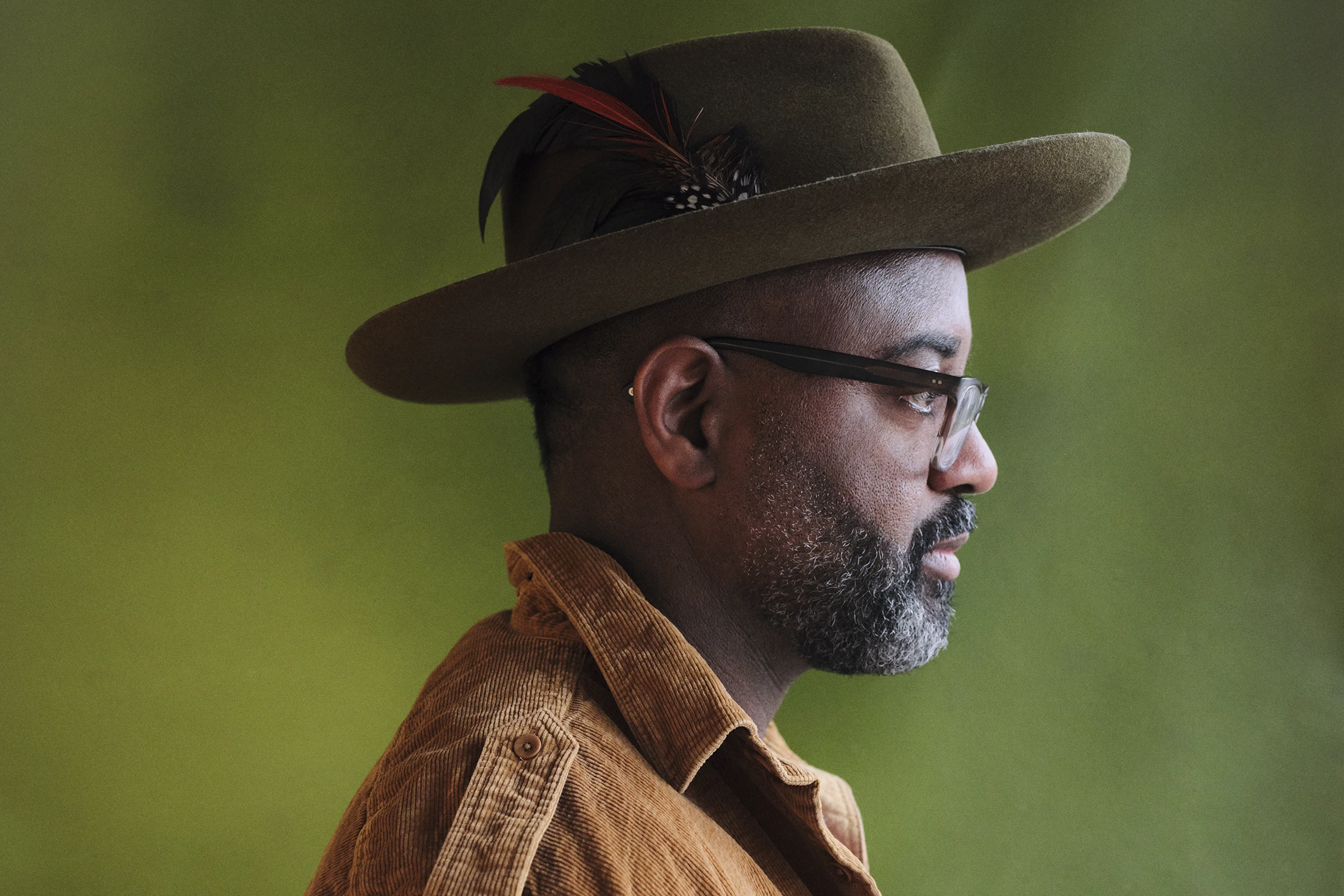 A side profile of a Black poet wearing glasses and a hat with a small feather hat pin. 