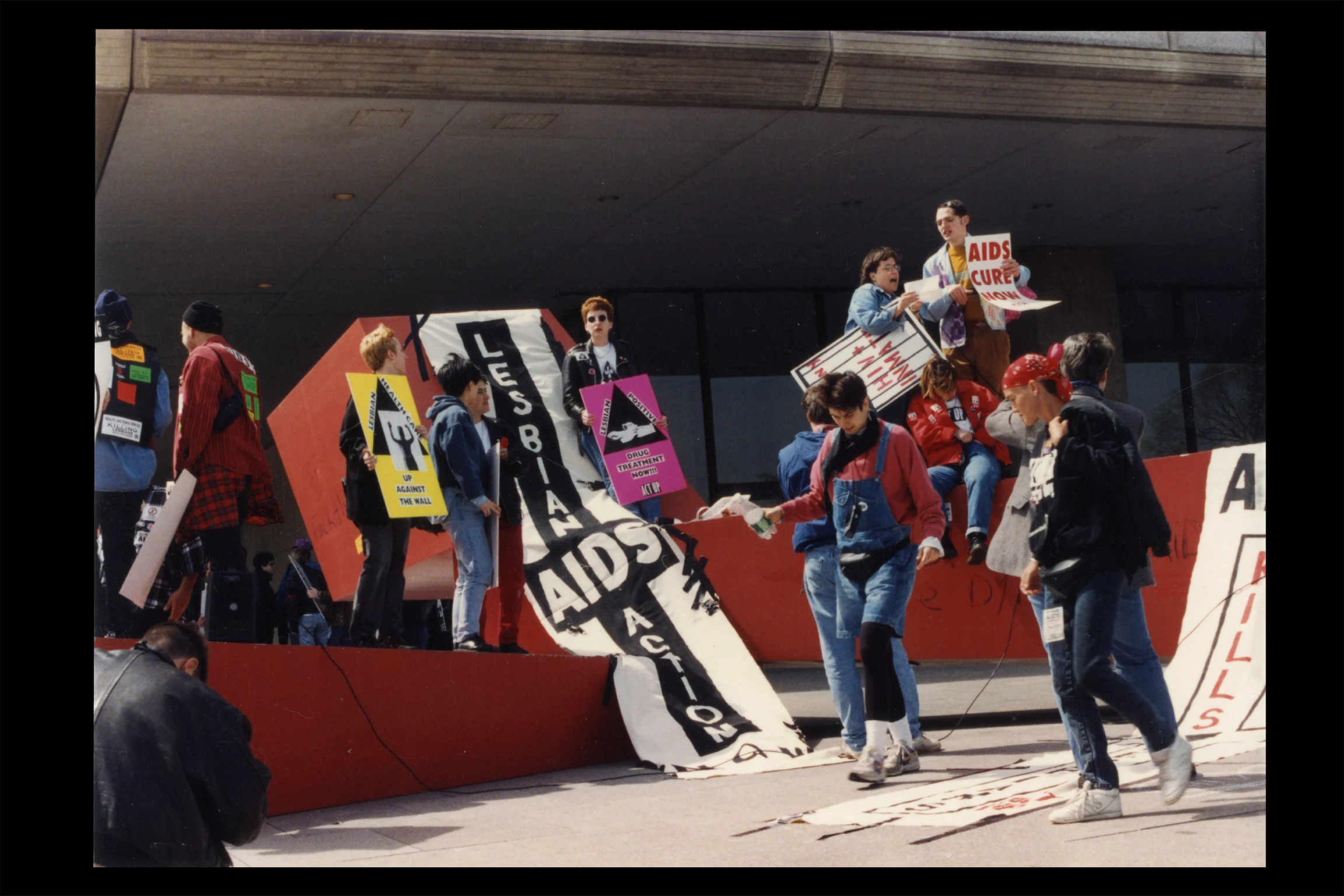 Color photograph of AIDS activists gathered on a stage during a protest, holding signs and banners with messages including “AIDS Cure Now!” and “Lesbian AIDS Action.”