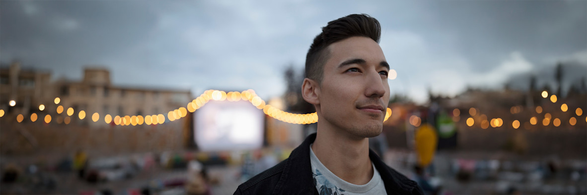 Un joven con cabello largo y fluido posa al aire libre bajo una cálida luz natural.