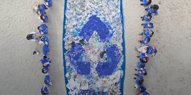 Dos filas de personas vistas desde arriba con camisetas azules y en un rectangulo en centro toda la basura recolectada con un simbolo de reciclado azul