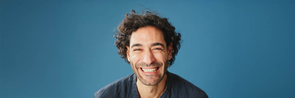 Un joven sonriente con cabello rizado natural está de pie contra un fondo azul.