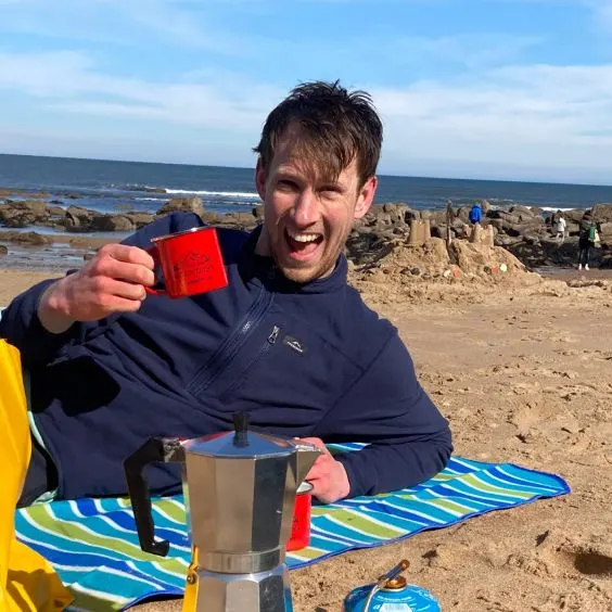 Man lying on Tynemouth beach with metal cup of coffee