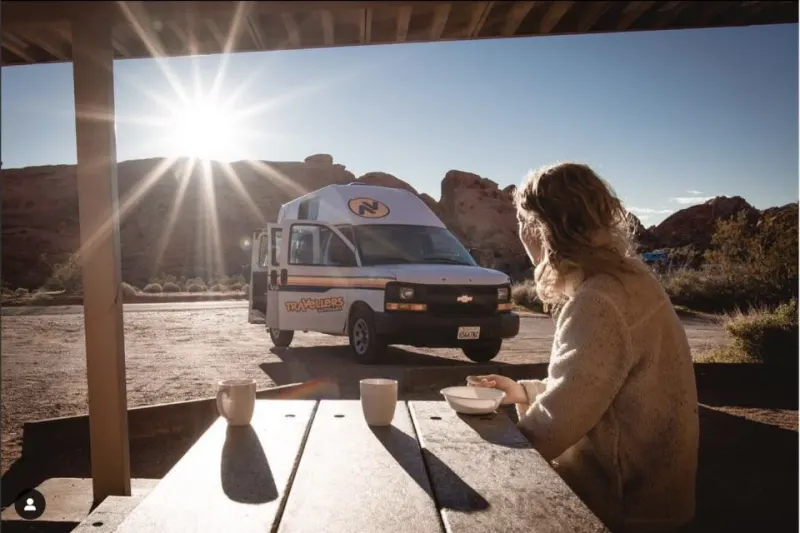 Woman sat on picnic bench in front of camper van in a desert with the sun shining brightly