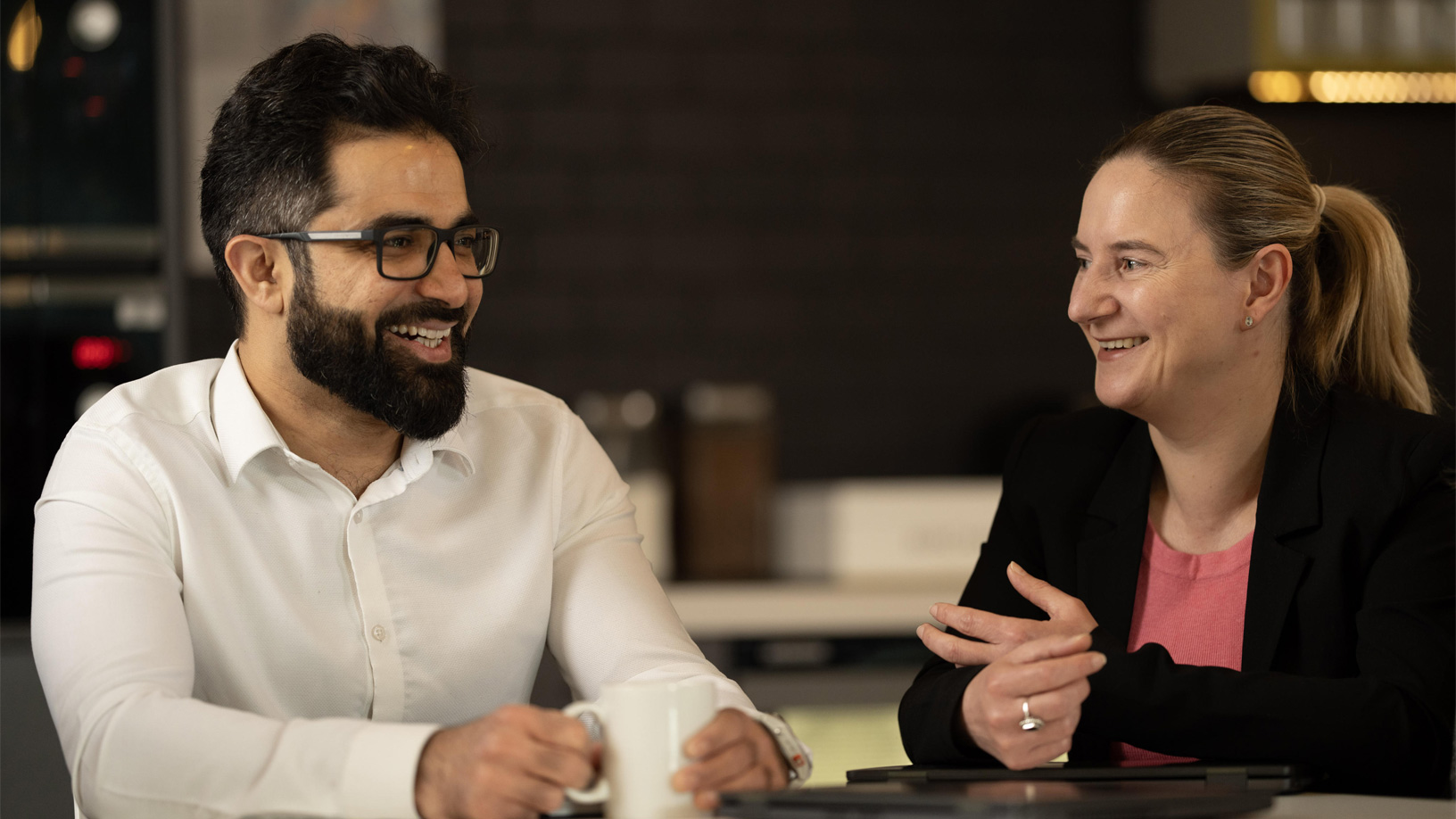 Two people smiling and talking at a table, one holding a mug. The setting is a warmly lit indoor space.