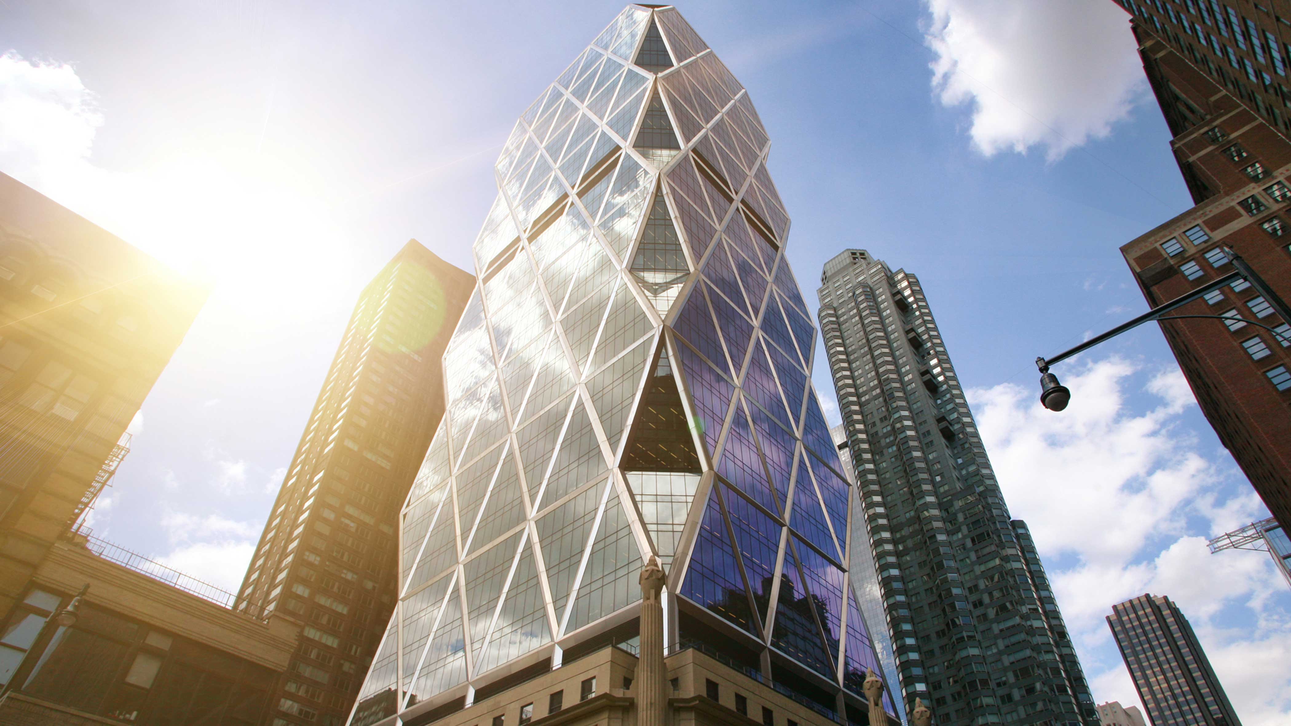 Low-angle view of a glass skyscraper with white diagonal grid, sun flare, and surrounding city towers under blue sky. on Media Contact